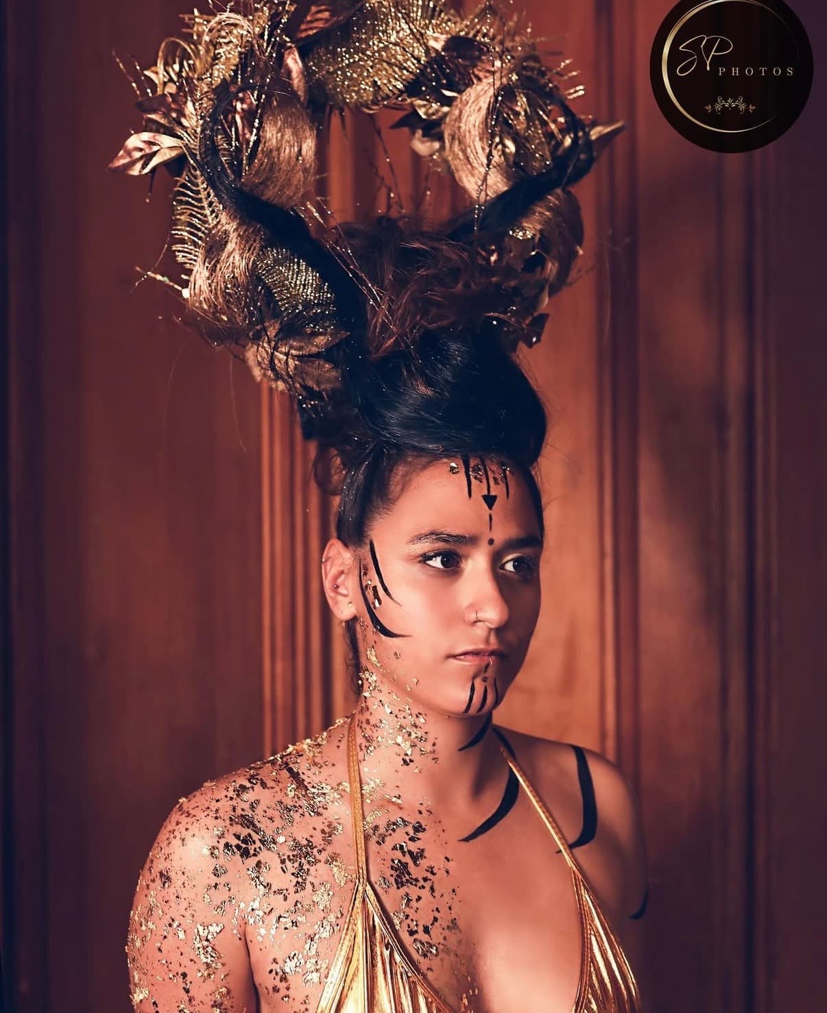 Un modèle avec une coiffure élaborée ornée d'éléments dorés et de marques faciales complexes, se tenant contre un fond en bois, capturant harmonieusement la beauté et la créativité.