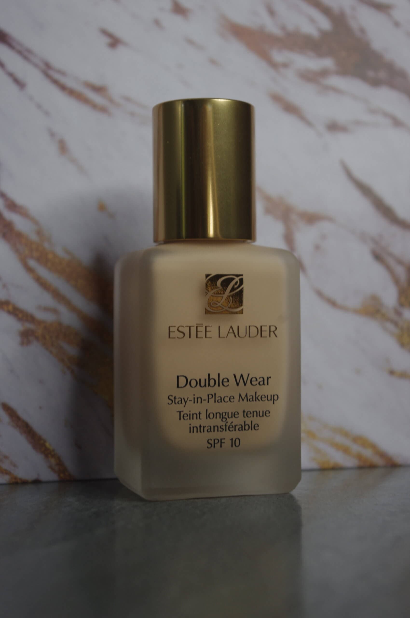 Un gros plan d'une bouteille de maquillage Estee Lauder Double Wear Stay-in-Place avec SPF 10, dotée d'un capuchon doré, sur fond marbré. L'étiquette affiche le nom du produit en anglais et en français, soulignant l'élégance de ce produit de beauté.