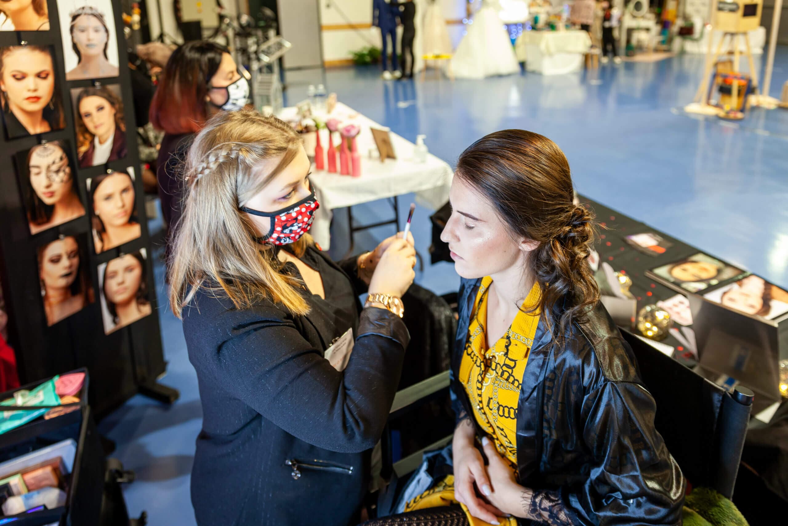 Une maquilleuse applique du maquillage à une cliente dans un cadre d'événement spacieux, alors que la cliente, portant un haut à motifs jaunes et les yeux fermés, reçoit une attention particulière. La maquilleuse, en masque et tenue élégante, se concentre sur le visage de la cliente. À l'arrière-plan, un panneau d'affichage présente un collage de différents looks de maquillage sur des modèles, entouré de tables avec des produits de beauté et des participants engagés dans l'événement, créant une atmosphère vibrante et professionnelle qui met en valeur l'art de la beauté.