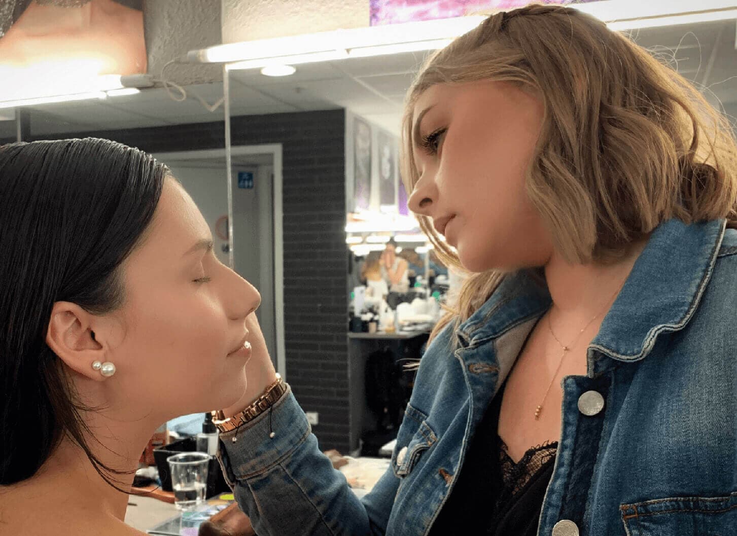 Un artiste maquilleur applique du maquillage à une femme aux longs cheveux noirs dans un salon de beauté, mettant en avant une expérience de service de beauté intime et attentive.