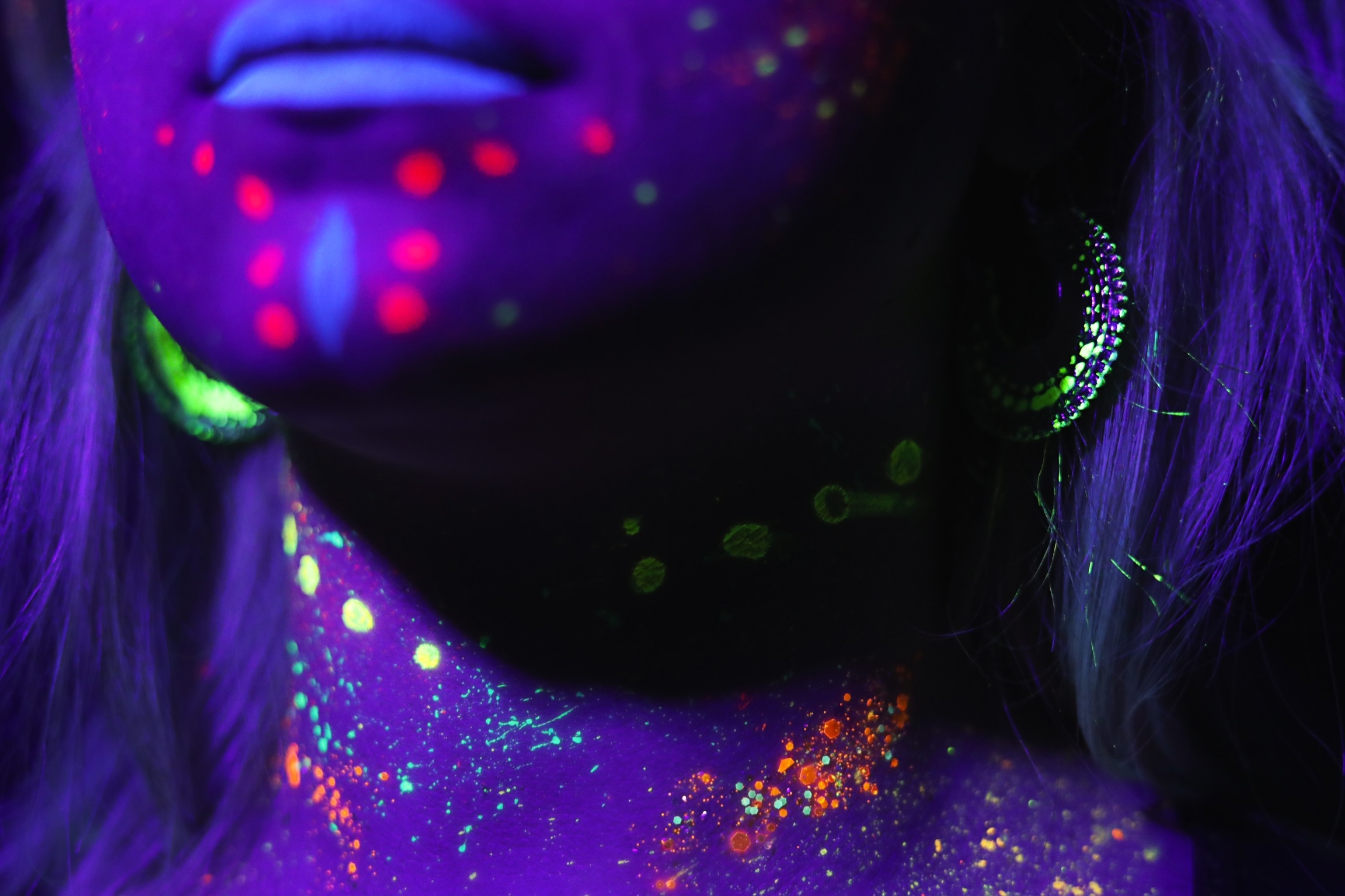 Gros plan sur le cou et le visage d'une personne illuminée par de la peinture fluorescente colorée sous une lumière UV, ornée de motifs vibrants en orange, vert et rouge, avec des boucles d'oreilles brillantes, le tout sur un fond sombre.