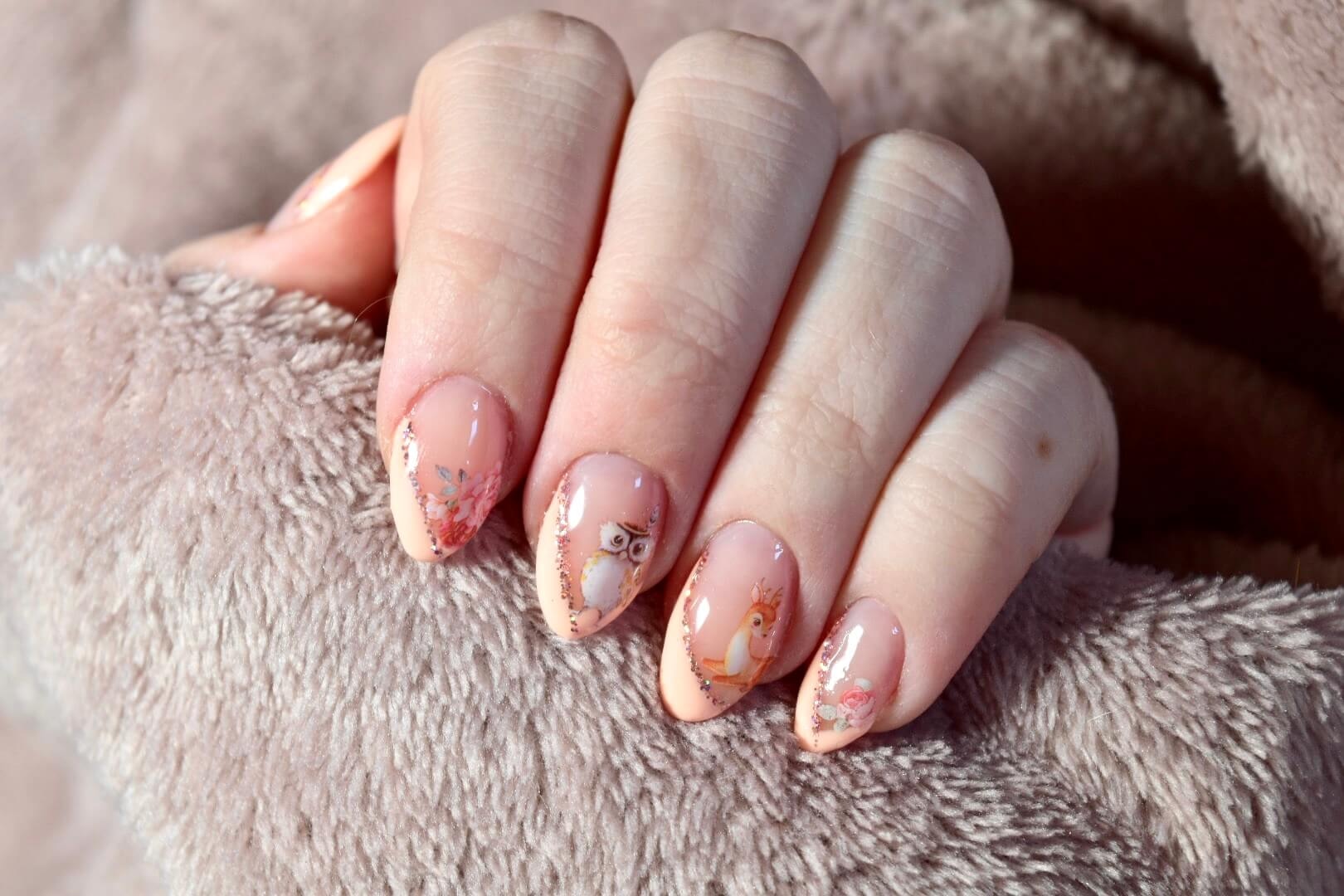 Gros plan sur une main posée sur une surface douce avec un vernis à ongles rose pâle, ornée de motifs délicats d'un hibou et d'éléments floraux, mettant en valeur l'élégance du design des ongles.