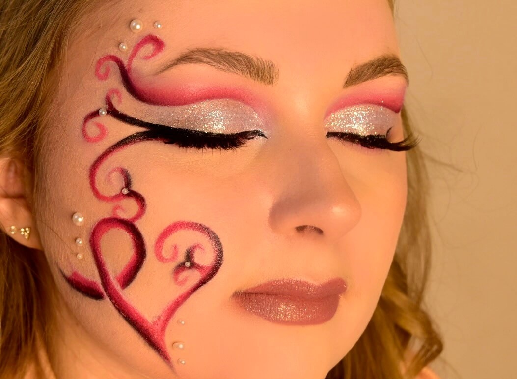 Gros plan sur une femme mettant en valeur un maquillage élaboré, avec une peau douce, des traits éclairés, et des lèvres rose clair. Son maquillage des yeux utilise des tons pastel et un argent scintillant, accentuant ses cils longs. Des tourbillons rouges et roses ornent le côté de son visage en forme de cœur, agrémentés de petites perles. L'arrière-plan est légèrement flou, soulignant les détails artistiques du maquillage.