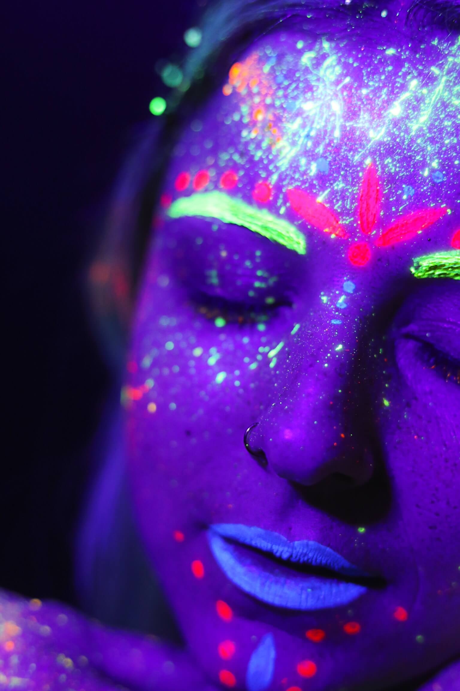 Un portrait en gros plan d'une personne aux yeux fermés, mettant en valeur des tatouages corporels vibrants sous la lumière ultraviolette, avec de la peinture néon colorée en vert, rose et bleu, sur fond sombre et violet.