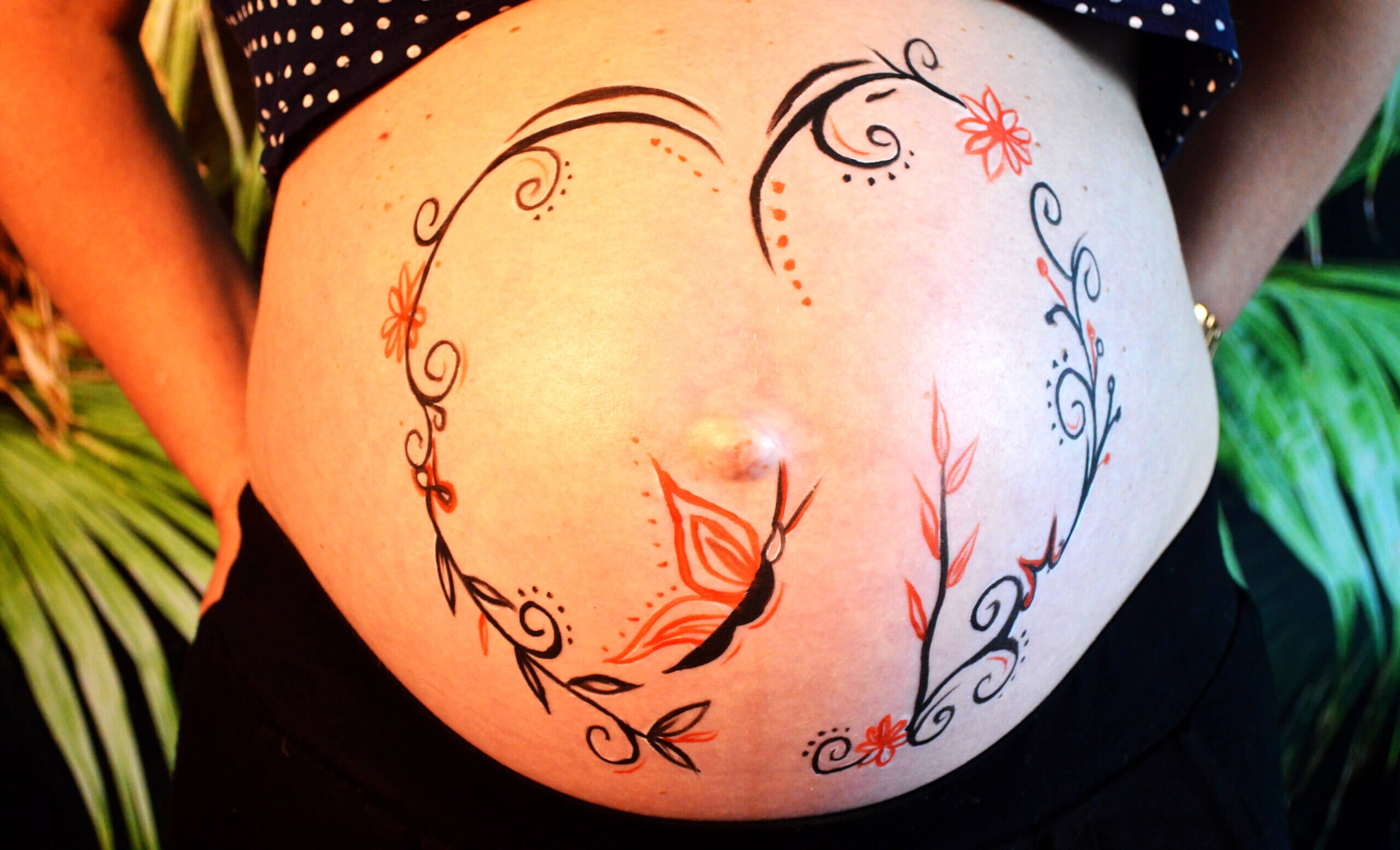 Gros plan d'un ventre de femme enceinte décoré avec des motifs artistiques complexes, incluant des lignes noires en spirale, des fleurs vibrantes et un papillon, le tout formant une forme de cœur, avec un arrière-plan de feuillage vert.
