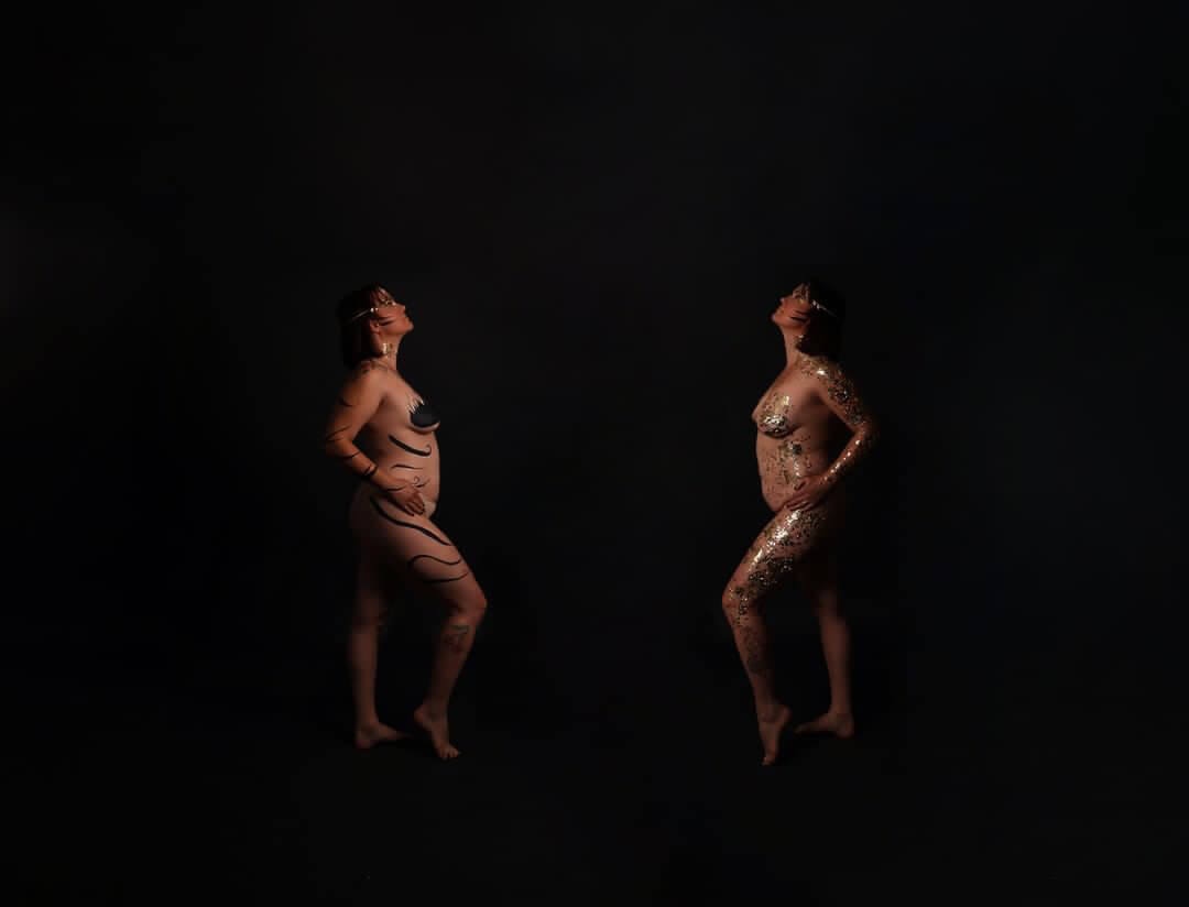 Deux figures debout sur un fond sombre, présentant des peintures corporelles artistiques avec des motifs ondulés sombres et de la peinture dorée scintillante.