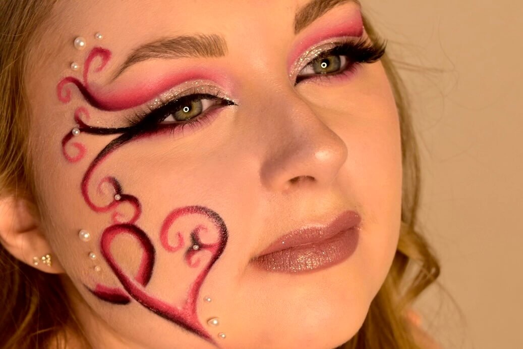 Gros plan sur une femme affichant un maquillage du visage élaboré, avec des motifs enroulés rouges et roses formant un cœur, accentués de petites perles blanches sur la joue.