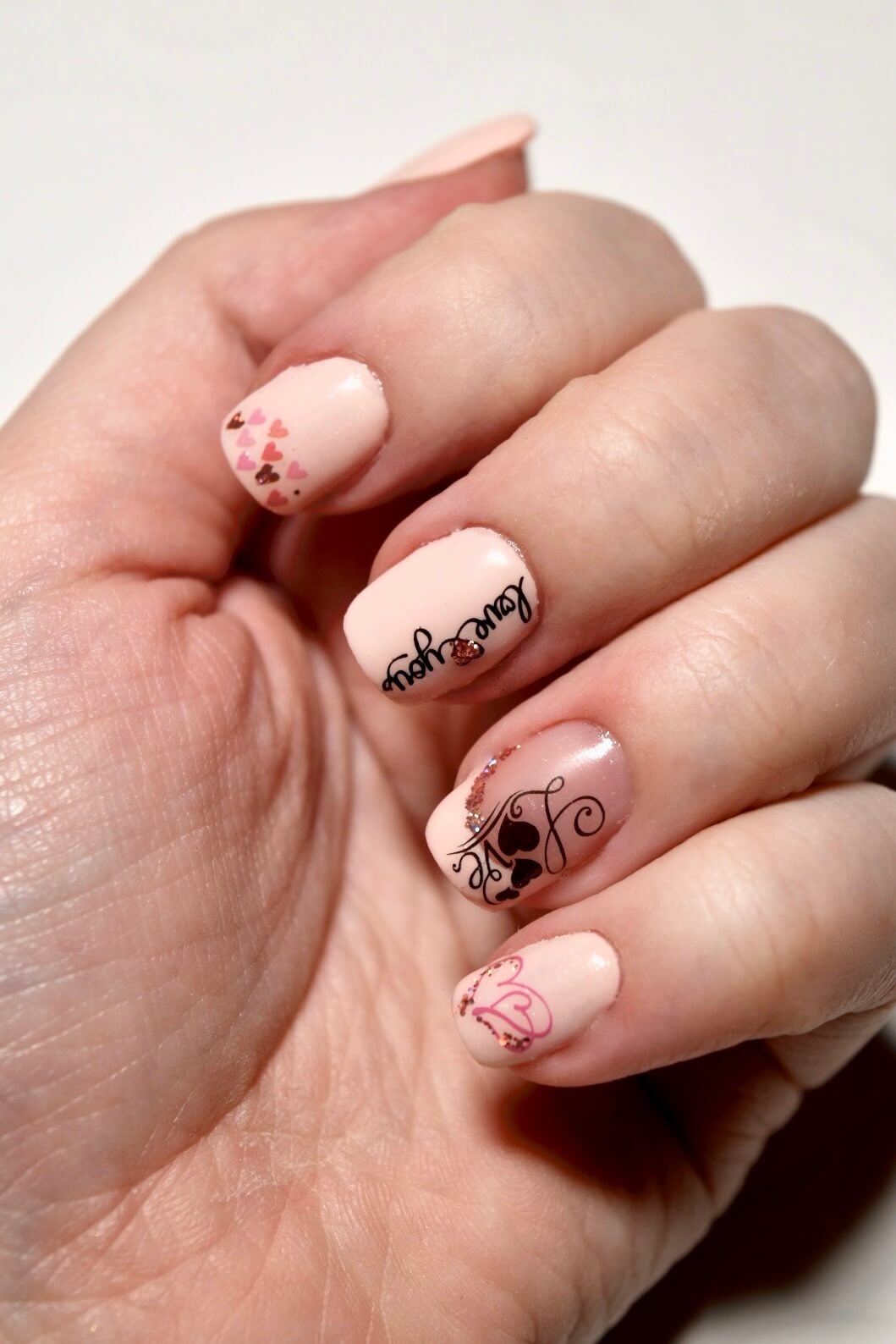 Gros plan d'une main affichant des ongles bien manucurés, avec une base rose clair ornée de petits cœurs rouges et roses, et un texte noir scripté qui dit 'Amour', le tout sur un fond blanc.
