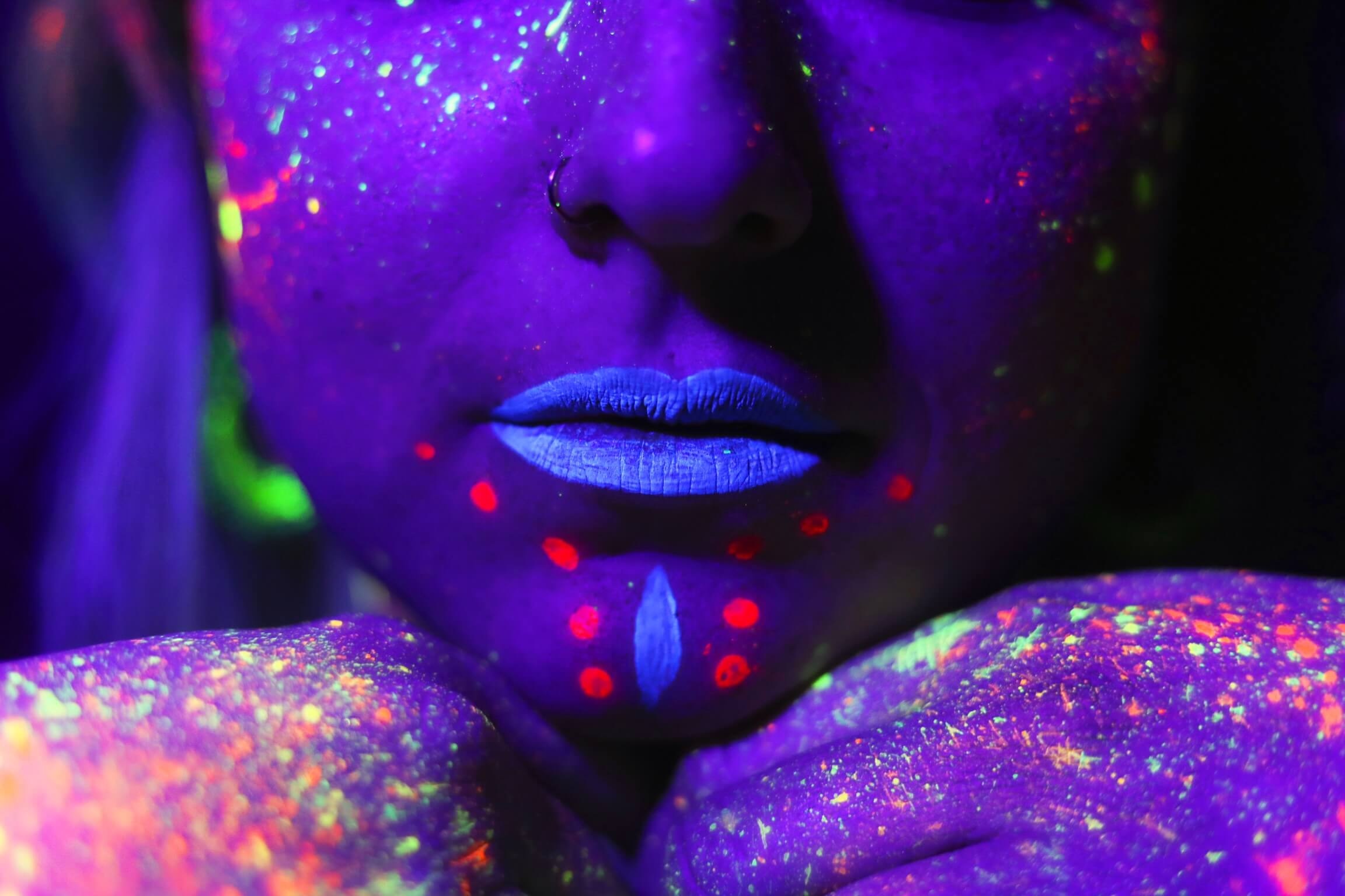 Premier plan d'un visage illuminé par des couleurs néon vives sous la lumière ultraviolette, avec des motifs brillants et des éclaboussures de peinture sur les joues, les lèvres et les épaules, créant un effet visuel saisissant.