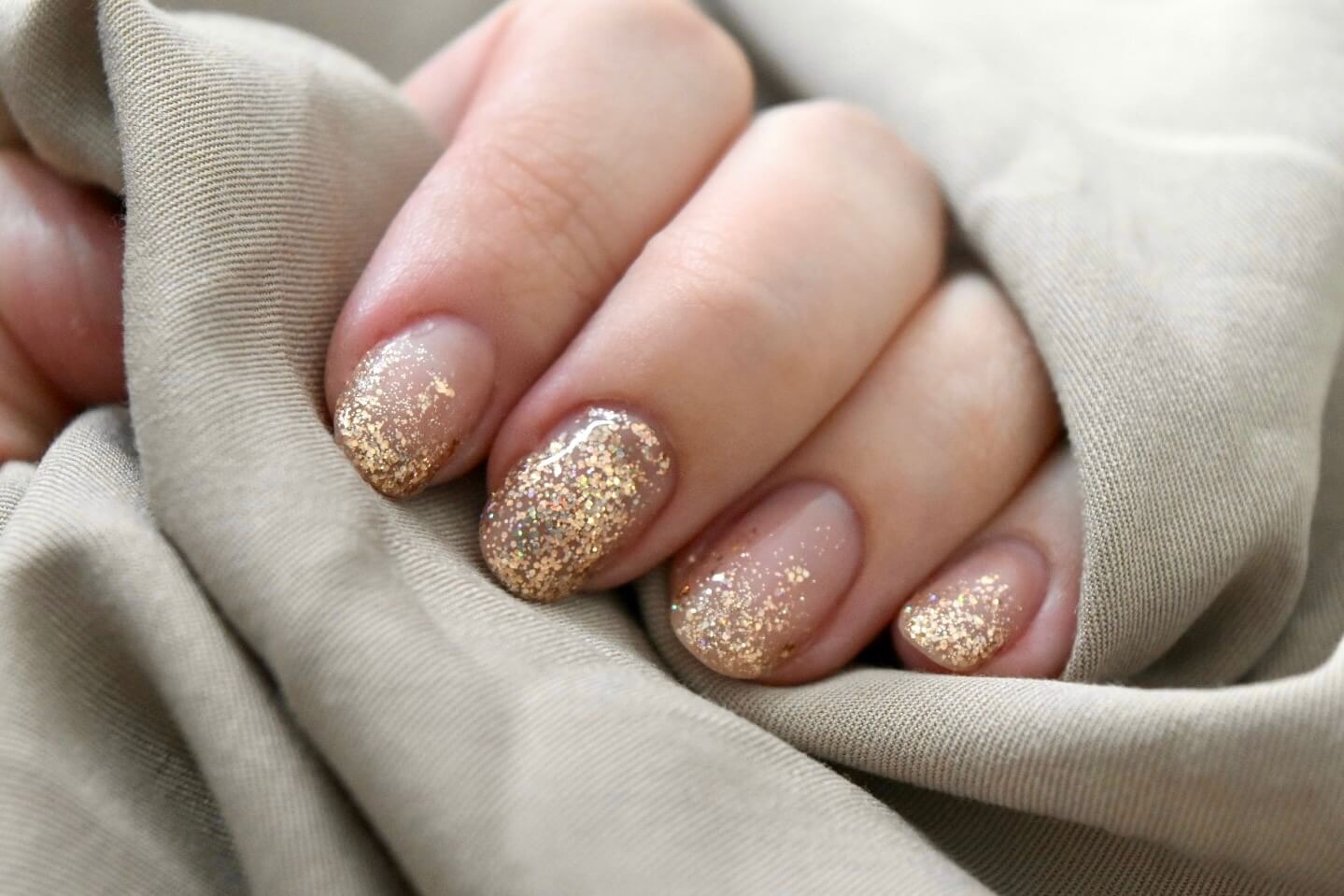 Gros plan d'une main reposant sur un tissu beige doux, mettant en valeur un vernis à ongles en dégradé nude et paillettes dorées, soulignant l'élégance de l'art des ongles moderne.