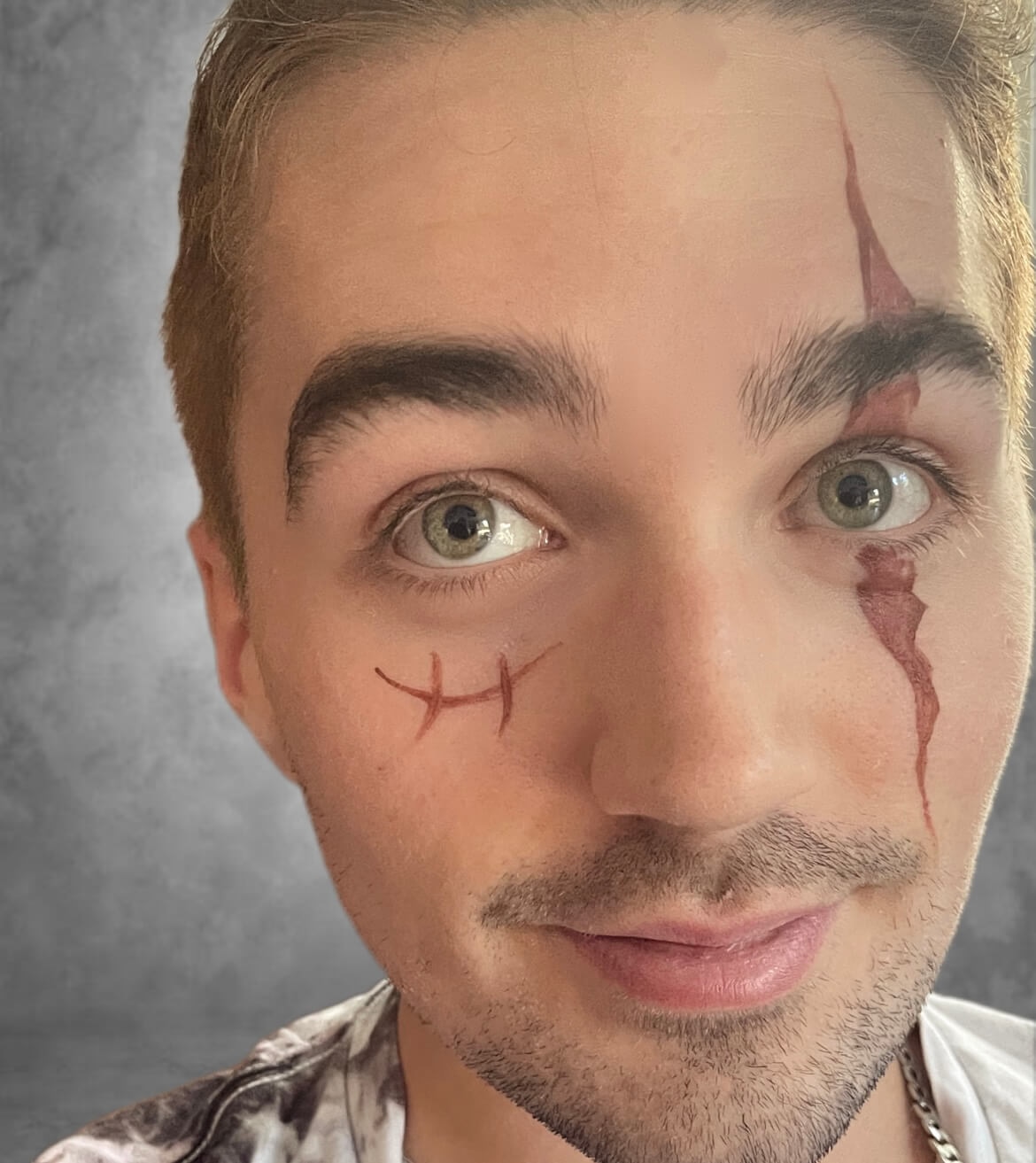 Un jeune homme souriant à l'objectif avec des designs de maquillage distinctifs, dont une ligne rouge en zigzag sur le visage et des lignes cousues sous l'œil, avec des cheveux châtain clair et des yeux verts, portant une chemise à motifs sur fond gris texturé.