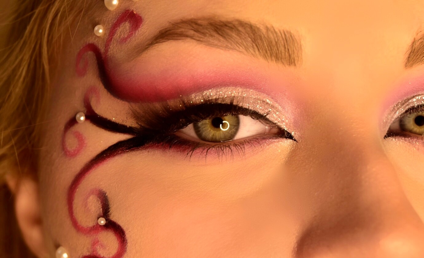 Gros plan sur l'œil d'une femme avec un maquillage artistique, mettant en valeur des nuances de rose vif et des paillettes argentées, entourées de tourbillons profonds et de petites perles, le tout sur un fond flou.