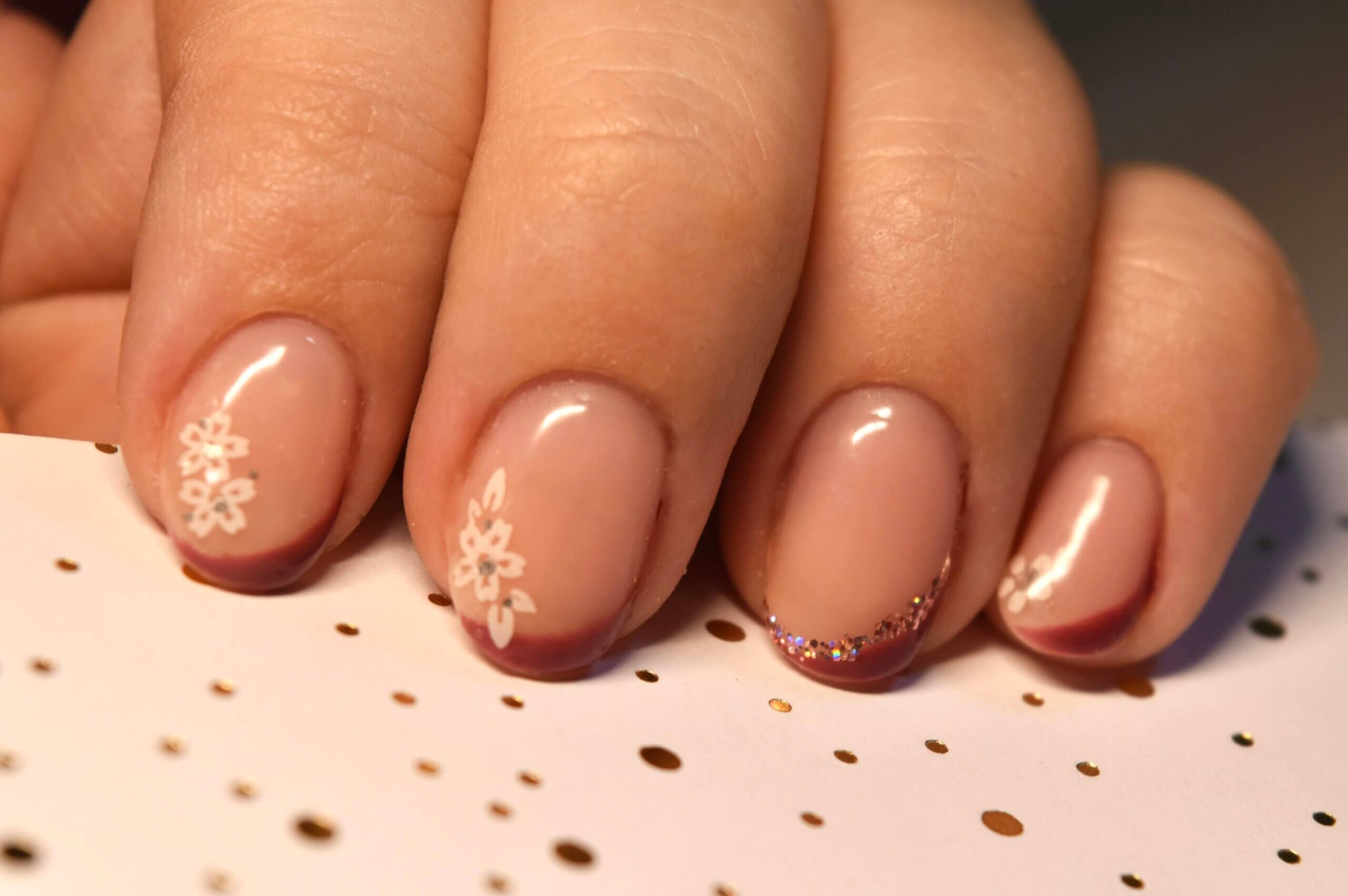 Une image en gros plan d'une main avec des ongles manucurés élégamment. Les ongles sont peints dans une nuance nude douce avec une finition brillante. Les annulaires et petits doigts présentent de délicats motifs floraux blancs, tandis que les bouts des ongles sont couleur bourgogne subtile. L'index est orné d'un accent scintillant avec des paillettes au bout. La main repose sur un fond texturé avec des pois dorés, ajoutant à l'esthétique générale de l'image.