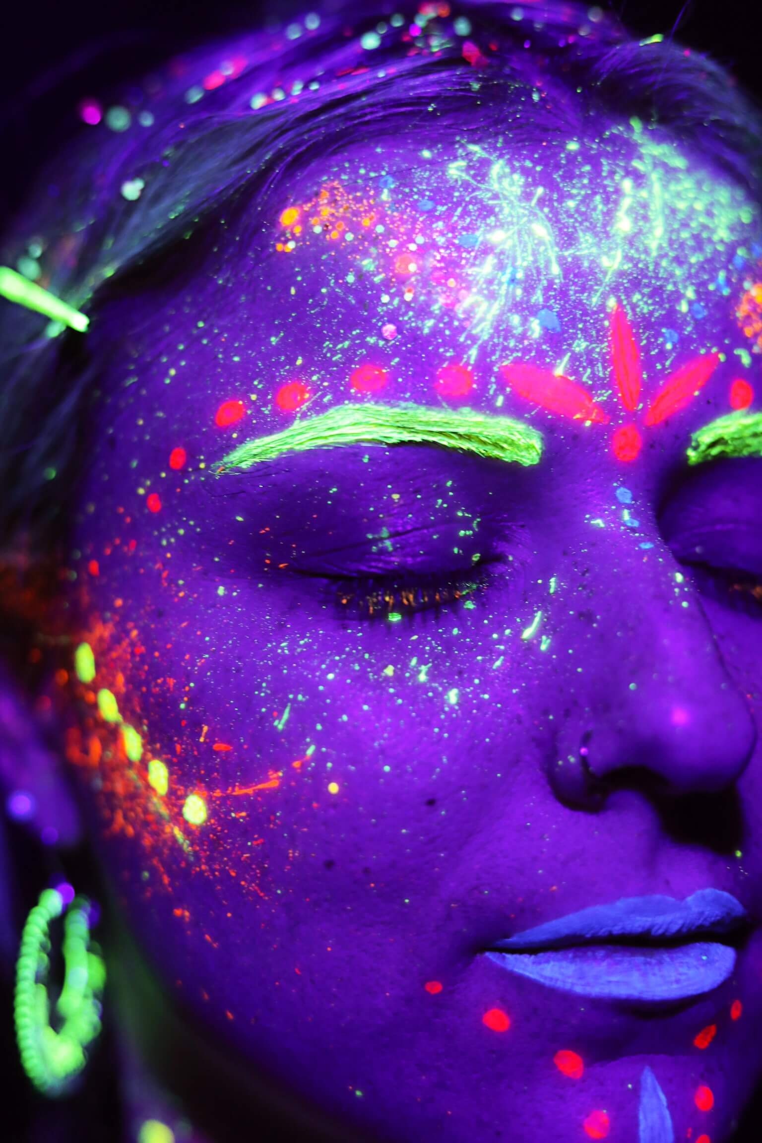 Gros plan sur le visage d'une personne illuminée par la lumière ultraviolette, ornée de motifs en peinture corporelle néon aux teintes vibrantes de bleu, rose et vert, créant un effet cosmique, avec les yeux fermés et des couleurs vives accentuant les sourcils et les lèvres.