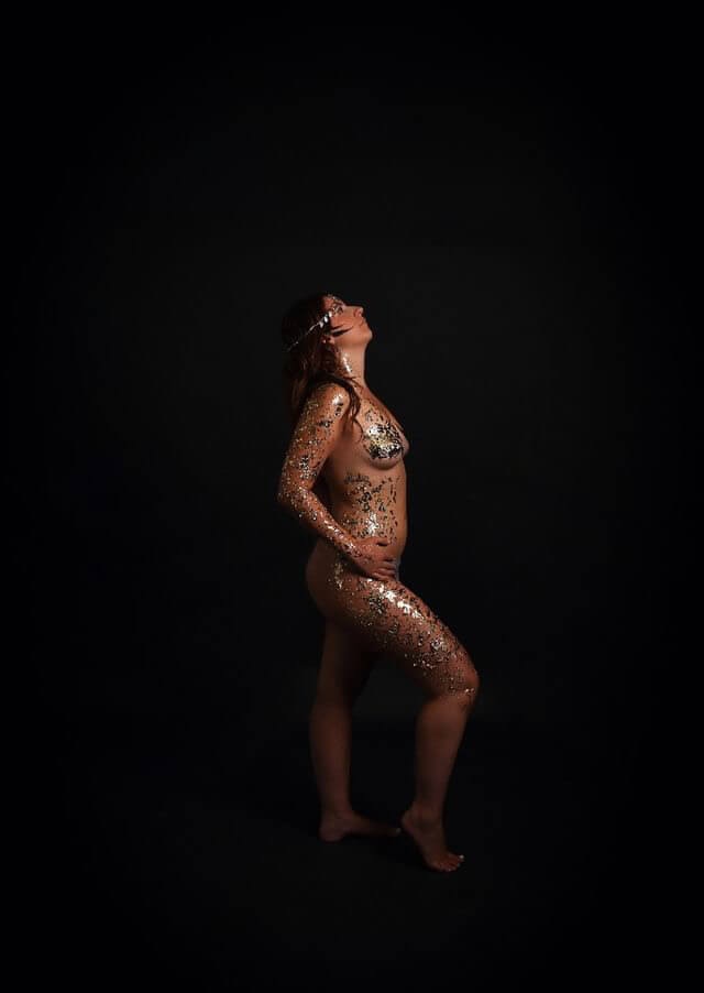 Une figure nue ornée de paillettes dorées pose en profil contre un fond sombre, regardant vers le haut avec une posture élégante, tandis que la lumière met en valeur les éclats et les contours de son corps, créant un contraste visuel saisissant.