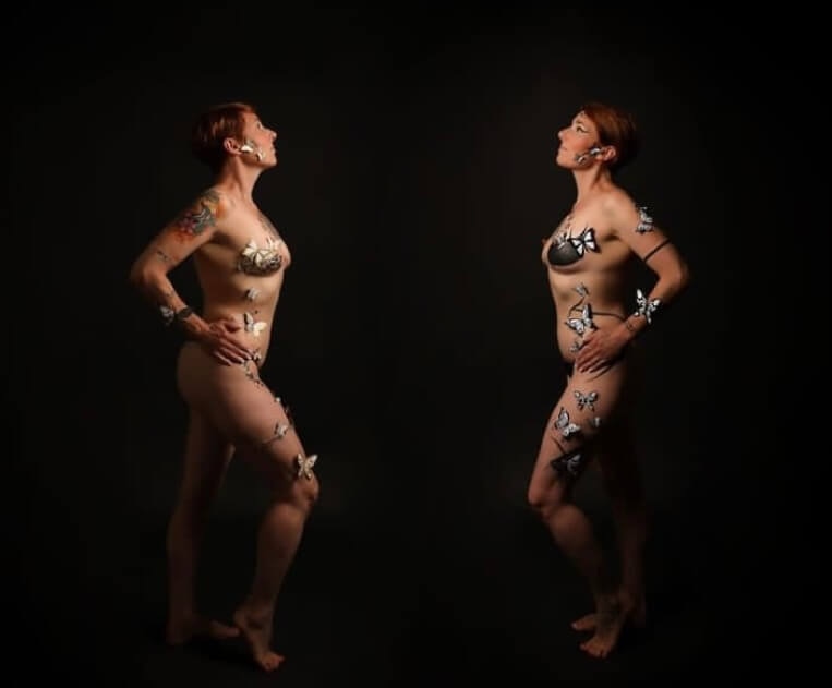 Deux femmes nues, dos à dos, ornées de motifs de papillons délicats sur leur peau, se détachent sur un fond sombre, créant une atmosphère artistique et éthérée.