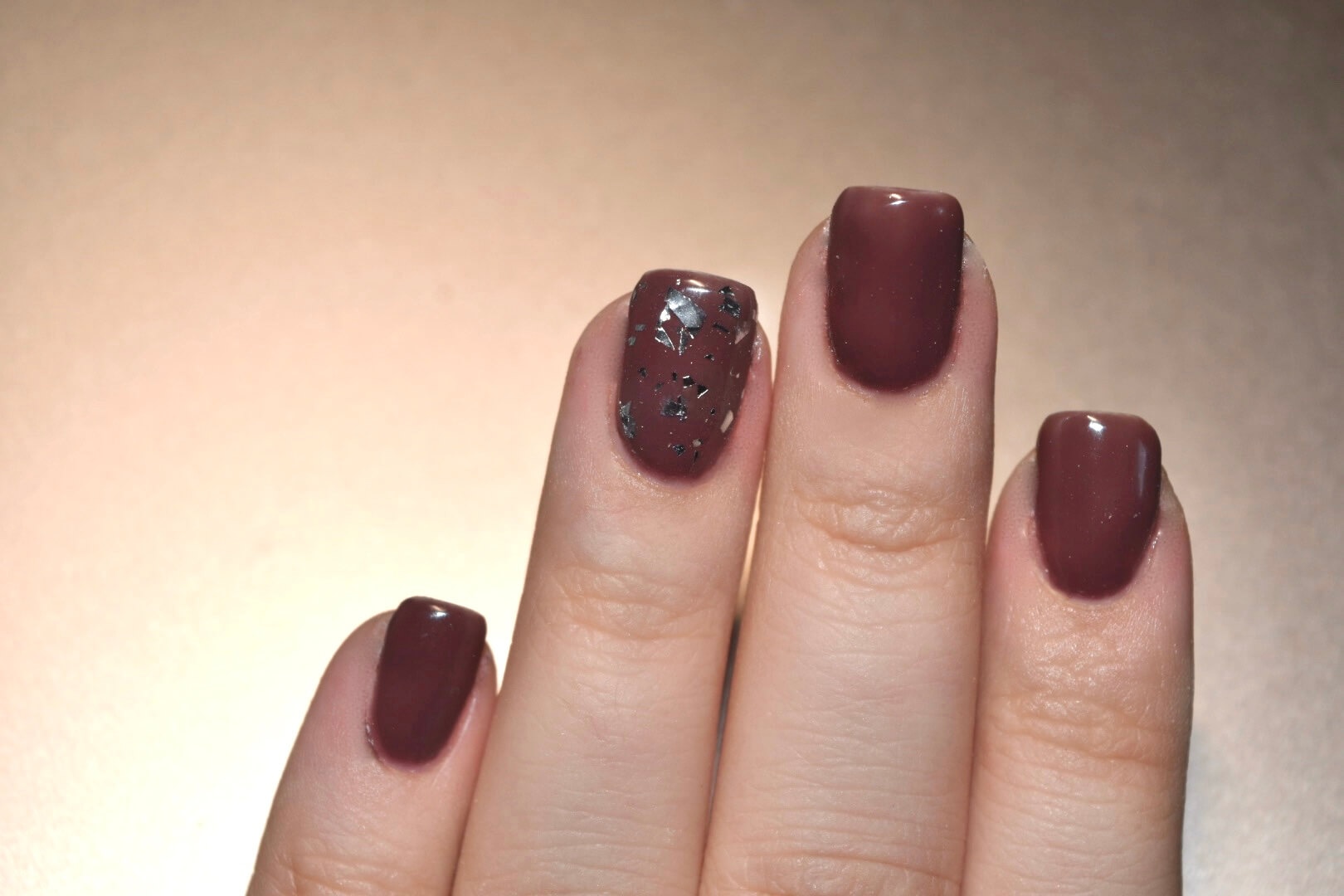 Gros plan sur une main manucurée avec des ongles peints en bourgogne, dont un présente un design décoratif avec des flocons métalliques argentés, le tout dans un éclairage doux et chaleureux.