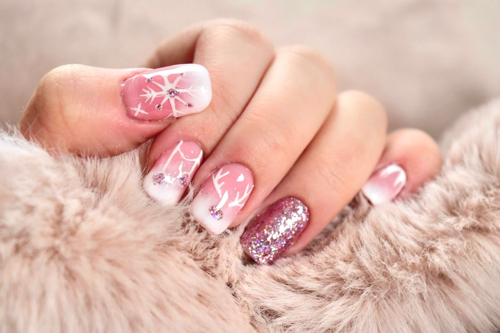 Gros plan d'une main avec des ongles manucurés, présentant un dégradé rose doux, des pointes blanches, des flocons de neige délicats et des embellissements scintillants, reposant sur une surface douce et confortable, parfait pour les occasions hivernales.