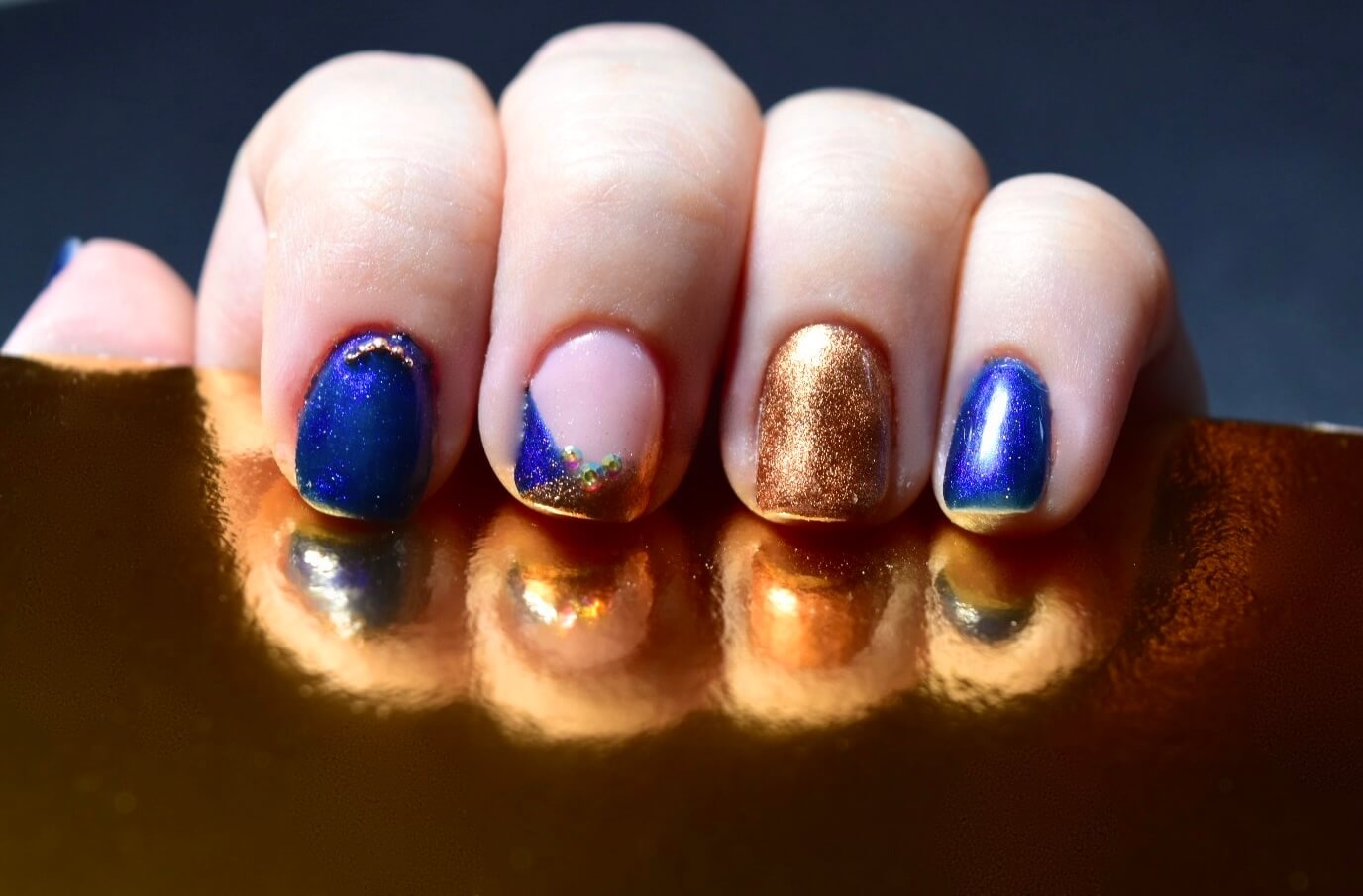 Une image en gros plan d'une main affichant des ongles manucurés avec une variété de couleurs de vernis à ongles, y compris un vernis bleu pailleté, une base nude avec des accents dorés, une finition brillante en bronze et un vernis métallique bleu profond, le tout sur un fond sombre qui met en valeur la beauté du design des ongles.