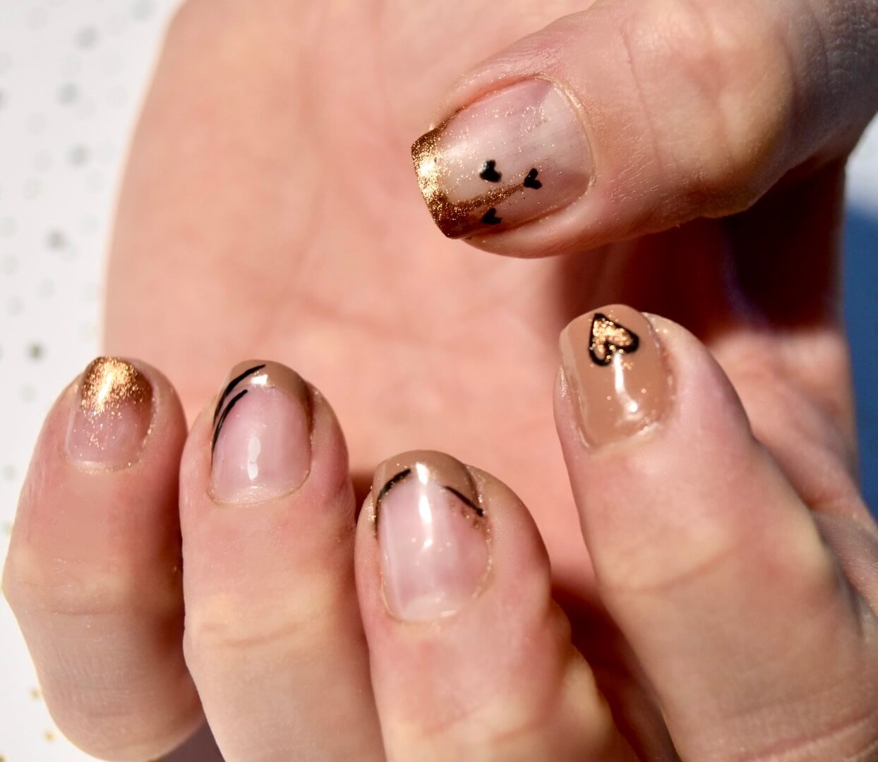 Gros plan d'une main mettant en valeur des ongles magnifiquement manucurés, ornés de vernis nude et d'accents dorés, avec des tips dorés et des designs de cœurs en noir sur certains ongles.