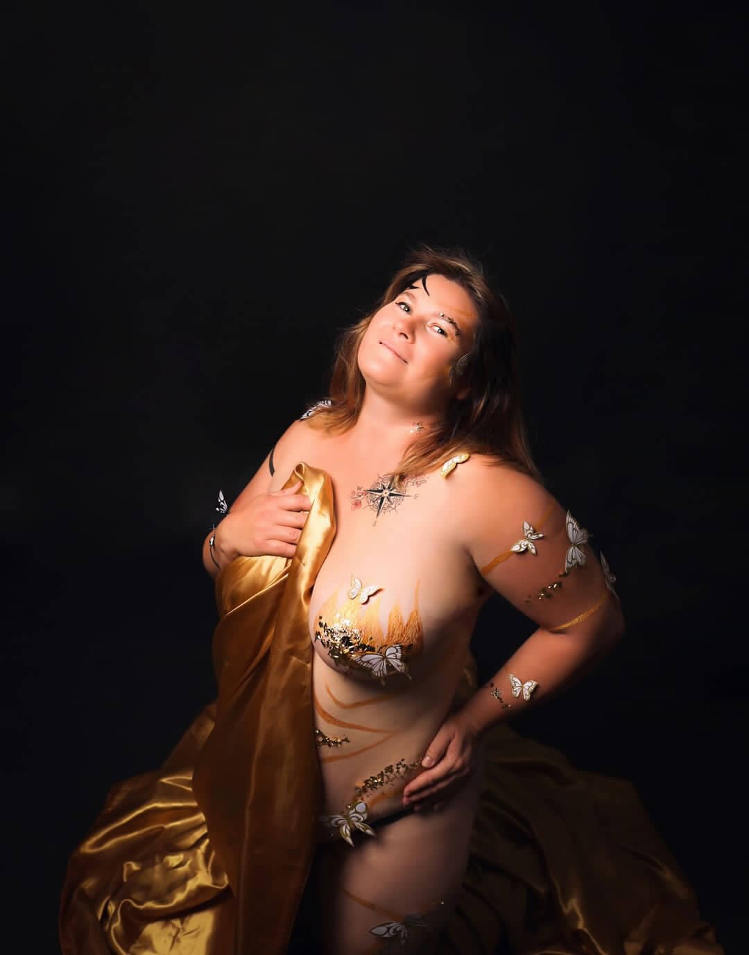 Une femme pose gracieusement dans un studio faiblement éclairé, entourée de peintures corporelles artistiques et d'éléments décoratifs comme des papillons et des motifs floraux, créant une connexion harmonieuse avec la nature.