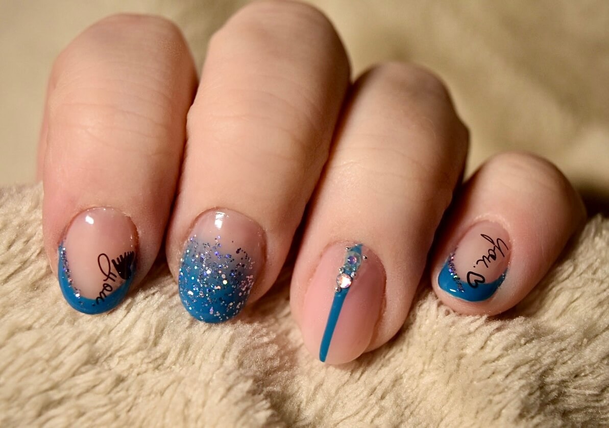 Manucure artistique avec un dégradé de vernis bleu et nude, présentant des designs délicats, des accents scintillants et un fond doux qui met en valeur les ongles.