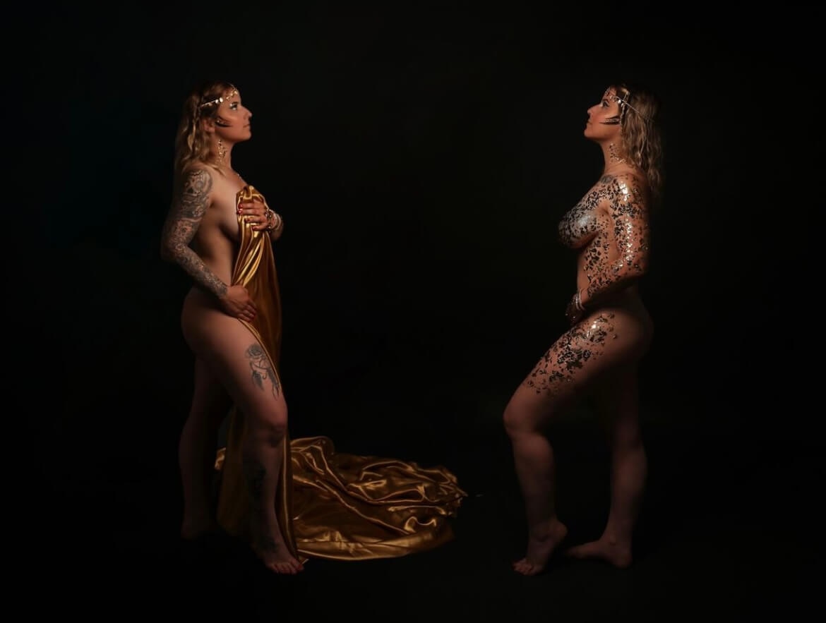 Deux femmes se tenant debout dans un environnement sombre ; l'une est drapée dans un tissu doré tandis que l'autre porte de la peinture corporelle artistique, mettant en valeur des tatouages et exprimant l'élégance.