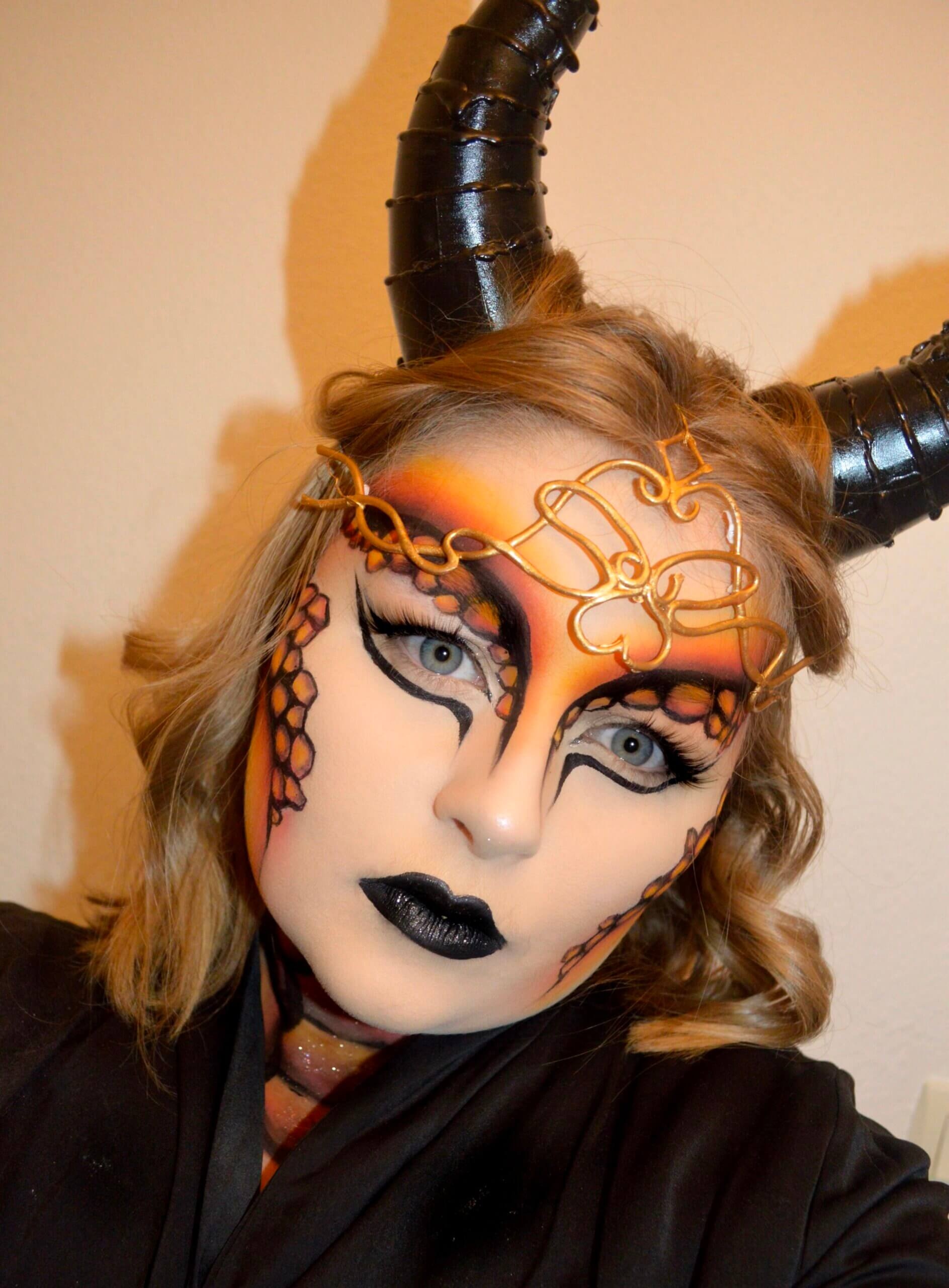 Portrait en gros plan d'une femme avec un maquillage dramatique et des accessoires, présentant des cornes noires spiralées et des peintures faciales orange et noires, dans une robe sombre.