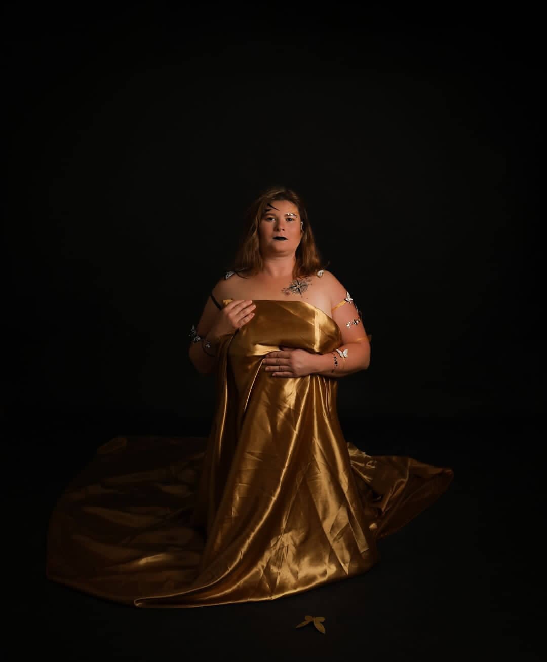 Une femme aux longs cheveux ondulés, assise avec grâce dans un cadre sombre, porte une élégante robe de satin doré ornée de détails argentés. Son maquillage audacieux aux lèvres noires souligne une ambiance contemplative alors qu'elle tient doucement ses mains, le fond noir mettant en valeur sa silhouette et la texture luxueuse de sa robe.