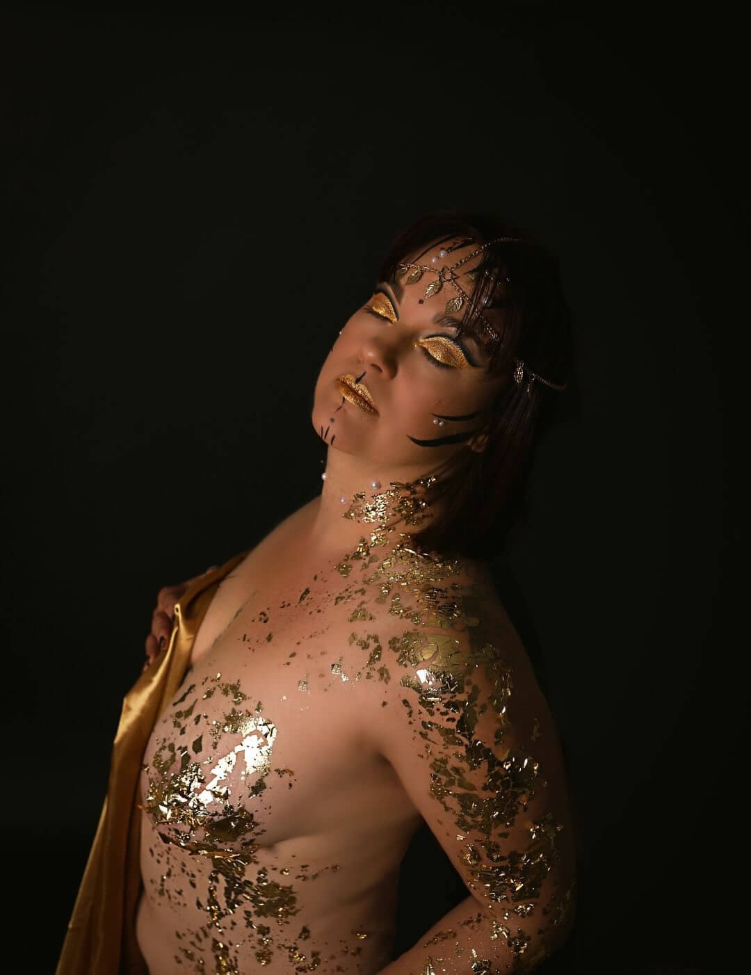 Un portrait en gros plan d'une personne ornée de maquillage doré complexe et d'art corporel, avec des flocons dorés scintillants sur leur visage et torse, créant une atmosphère de beauté et d'élégance.