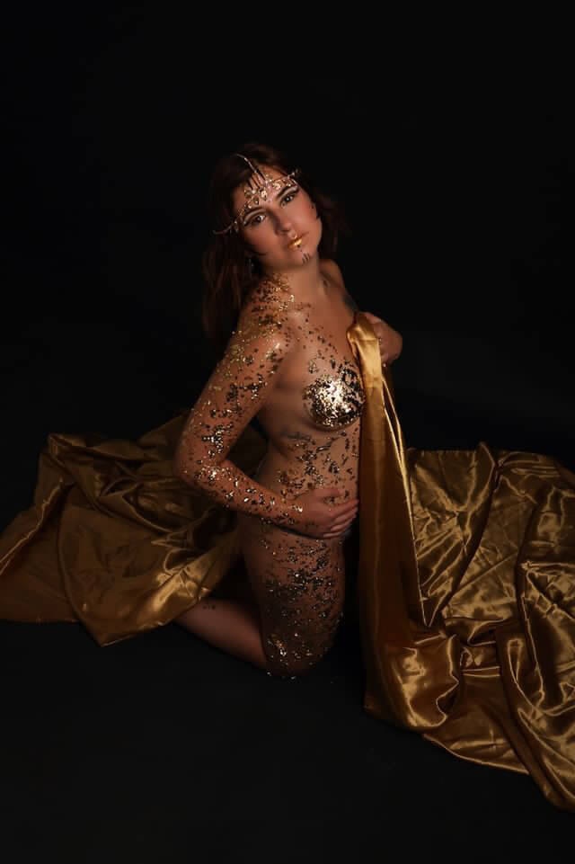 Une femme pose dans une photographie artistique sur fond noir, ornée de brillants embellissements corporels dorés et argentés qui scintillent. Un tissu satiné doré drape élégamment autour d'elle, tandis que ses cheveux cascades complètent le thème doré. Son expression est sereine et contemplative, et l'éclairage dramatique met en valeur ses traits et les détails complexes des ornements et du tissu.