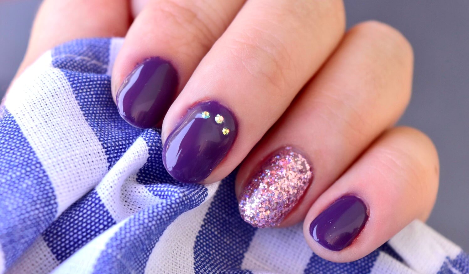 Gros plan sur une main avec des ongles manucurés de couleur violet foncé, un ongle décoré de paillettes roses et deux petits rhinestones, reposant sur un tissu rayé bleu et blanc avec un fond gris doux.