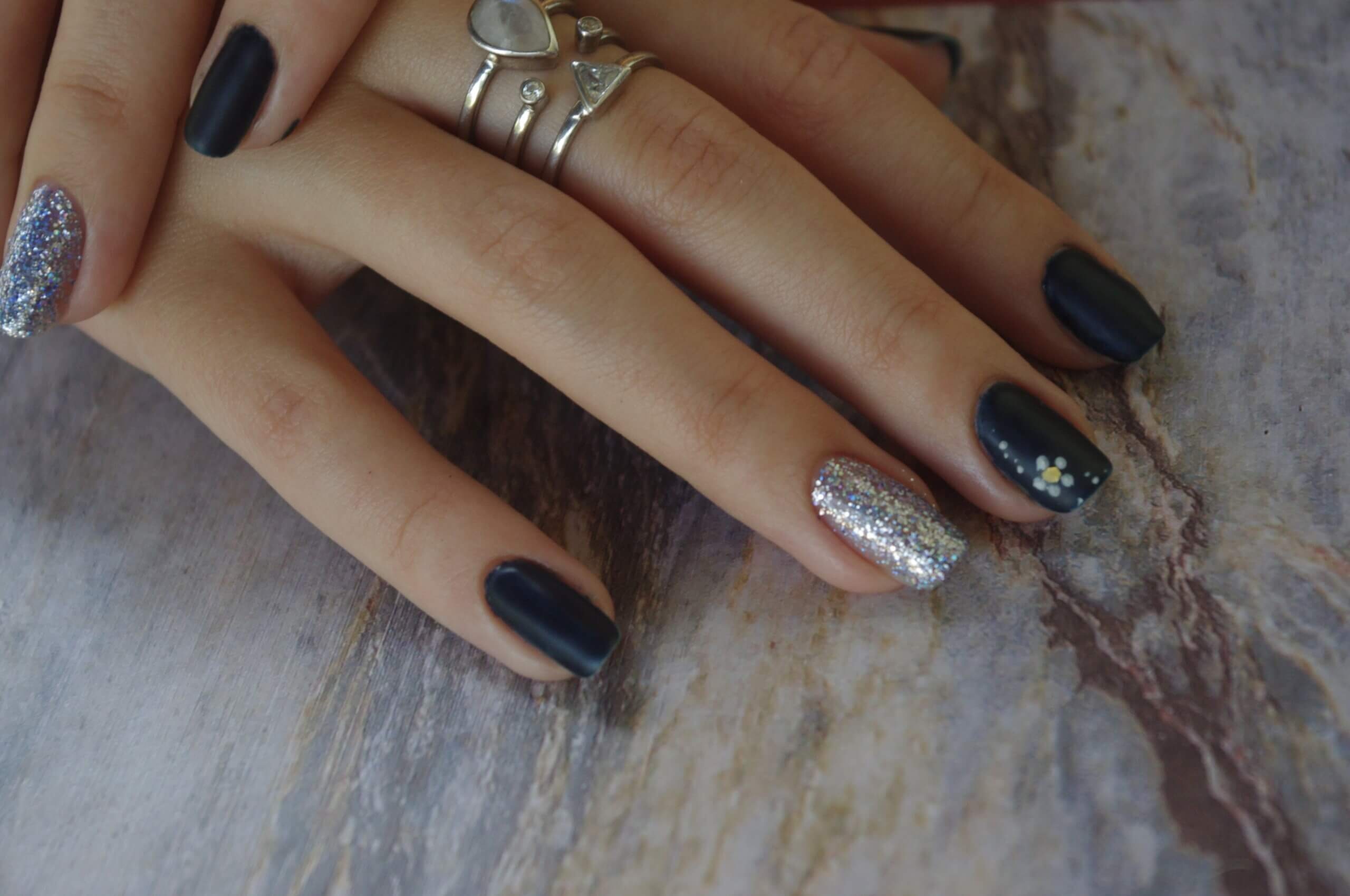 Gros plan sur une main reposant sur une surface texturée, montrant une manucure élégante avec des ongles peints en bleu marine mat et un ongle accent en paillettes argentées, décoré de petites fleurs blanches et jaunes, accompagnée de délicates bagues en argent.