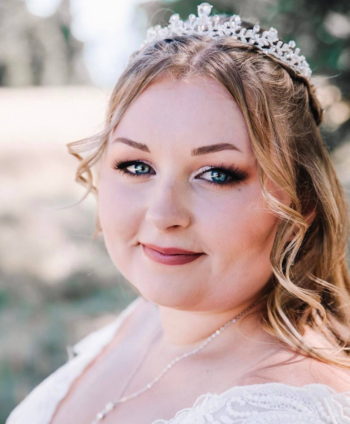 Un portrait rapproché d'une femme aux cheveux blonds et ondulés portant une délicate couronne, avec des yeux bleus saisissants et un maquillage subtil, vêtue d'une robe de mariée blanche ornée de dentelle, dans un cadre naturel flou.