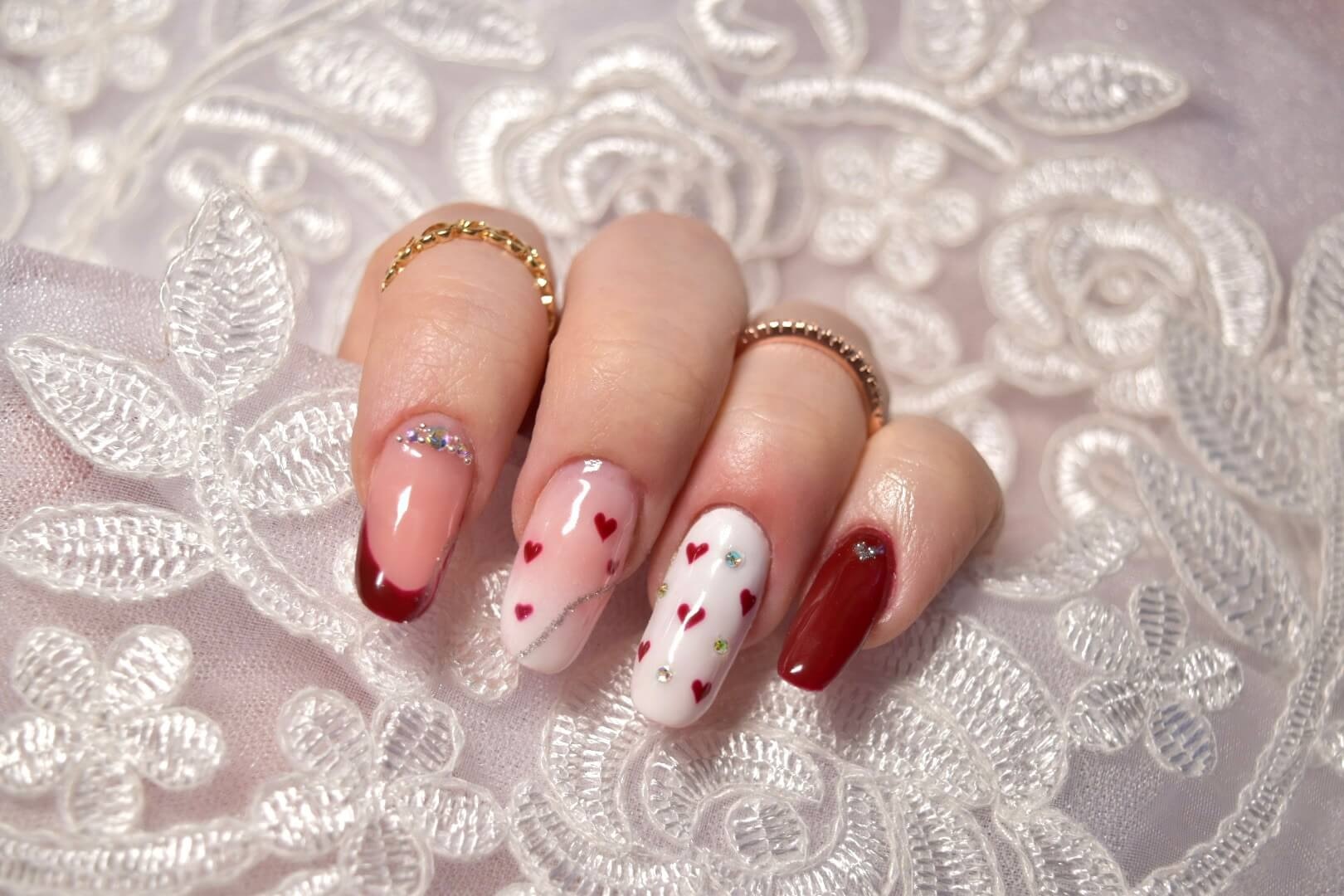Gros plan d'une main bien manucurée avec des ongles ornés de nail art, des finitions rouge profond et une base rose clair décorée de petits cœurs rouges et de gemmes colorées, posée sur un fond délicat en dentelle blanche.