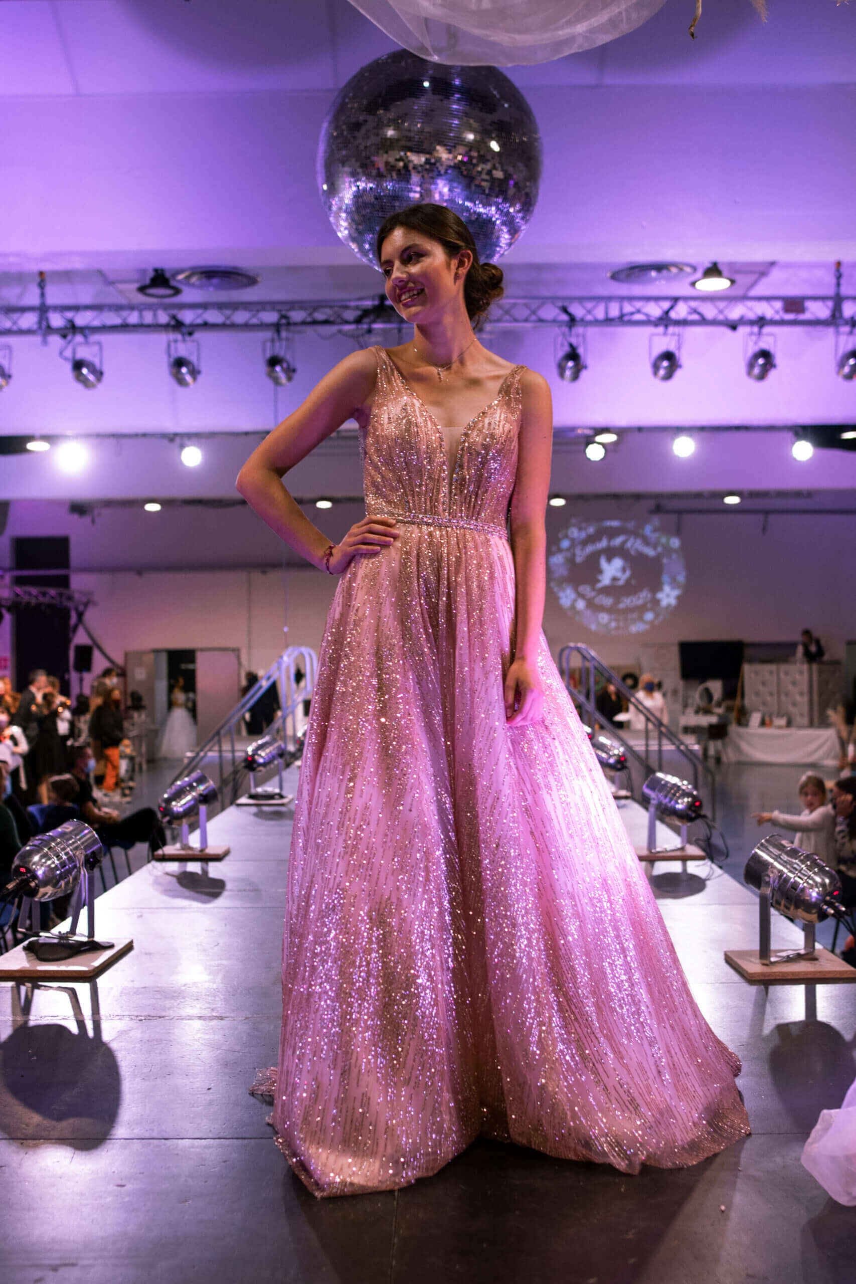 Un mannequin se tient sur un podium de mode, mettant en valeur une magnifique robe rose ornée de sequins qui scintillent sous les lumières, tandis qu'un grand miroir disco reflète la lumière et crée une atmosphère festive.