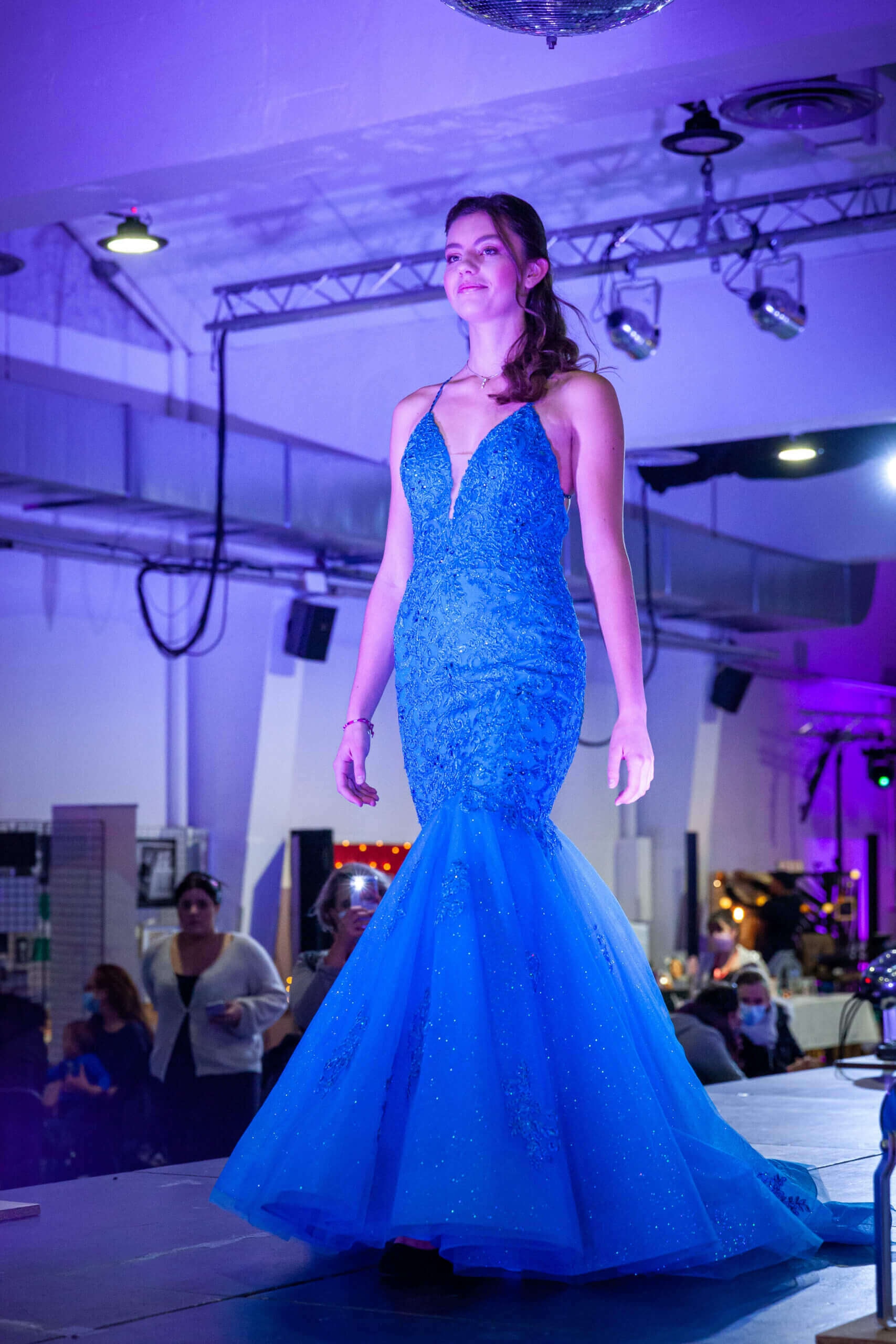 Une mannequin présente une robe bleue éblouissante avec un corsage ajusté et une jupe évasée sur un podium, ornée de embellissements scintillants sous les lumières, tandis qu'une ambiance stylée est créée par un éclairage violet et blanc en arrière-plan.
