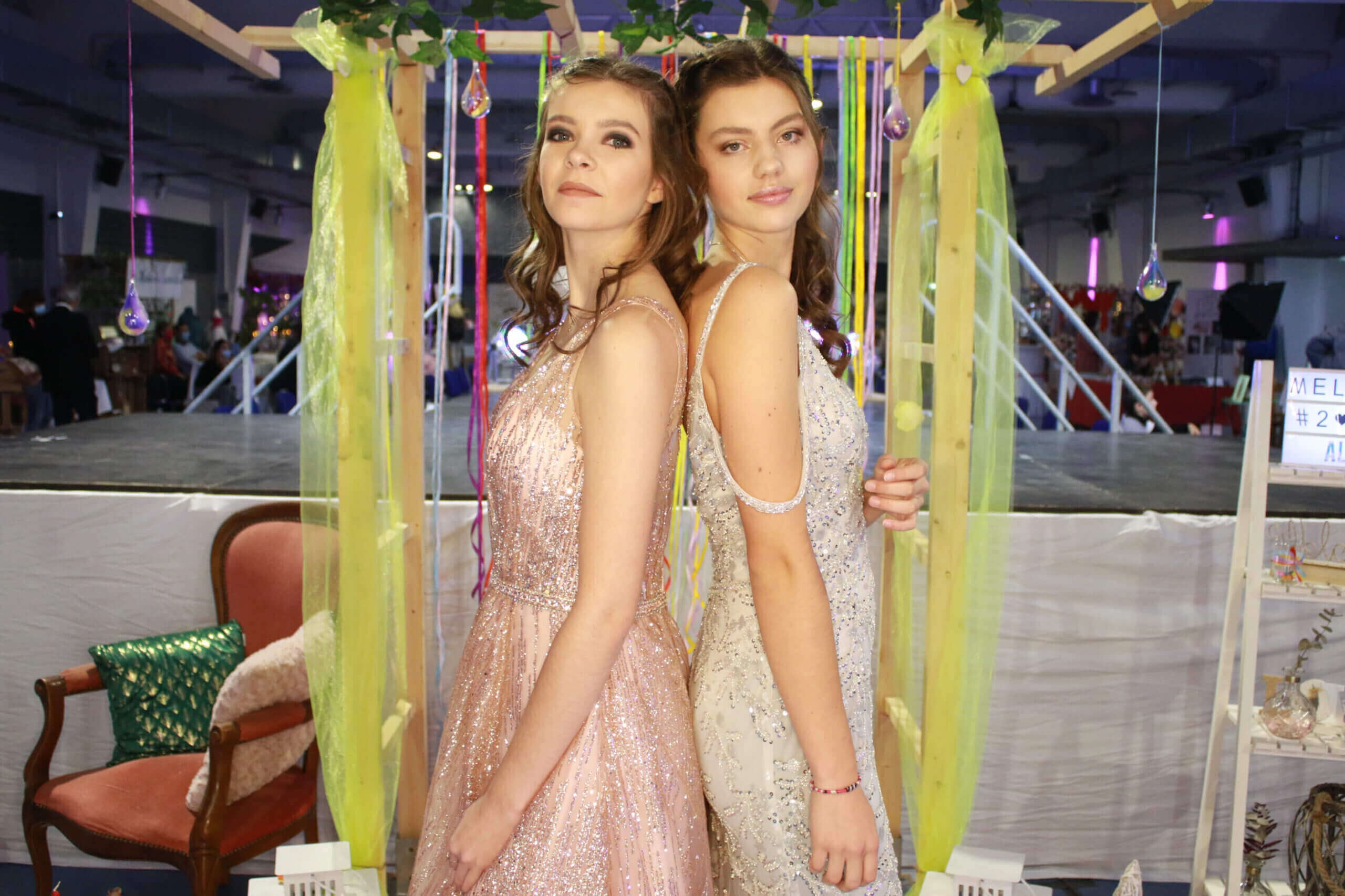 Deux mannequins posent dos à dos dans un cadre festif, l'un portant une robe rose scintillante et l'autre une robe argentée brillante, devant un décor de banderoles colorées et d'ornements en verre.