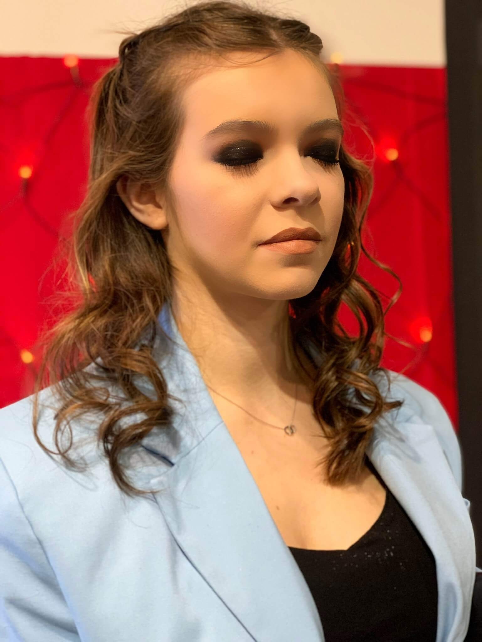 Une jeune femme aux cheveux bouclés porte un blazer bleu clair par-dessus un top noir, les yeux fermés et un maquillage dramatiquement foncé autour des yeux, se tenant dans une ambiance vibrante avec des lumières douces sur un fond rouge.