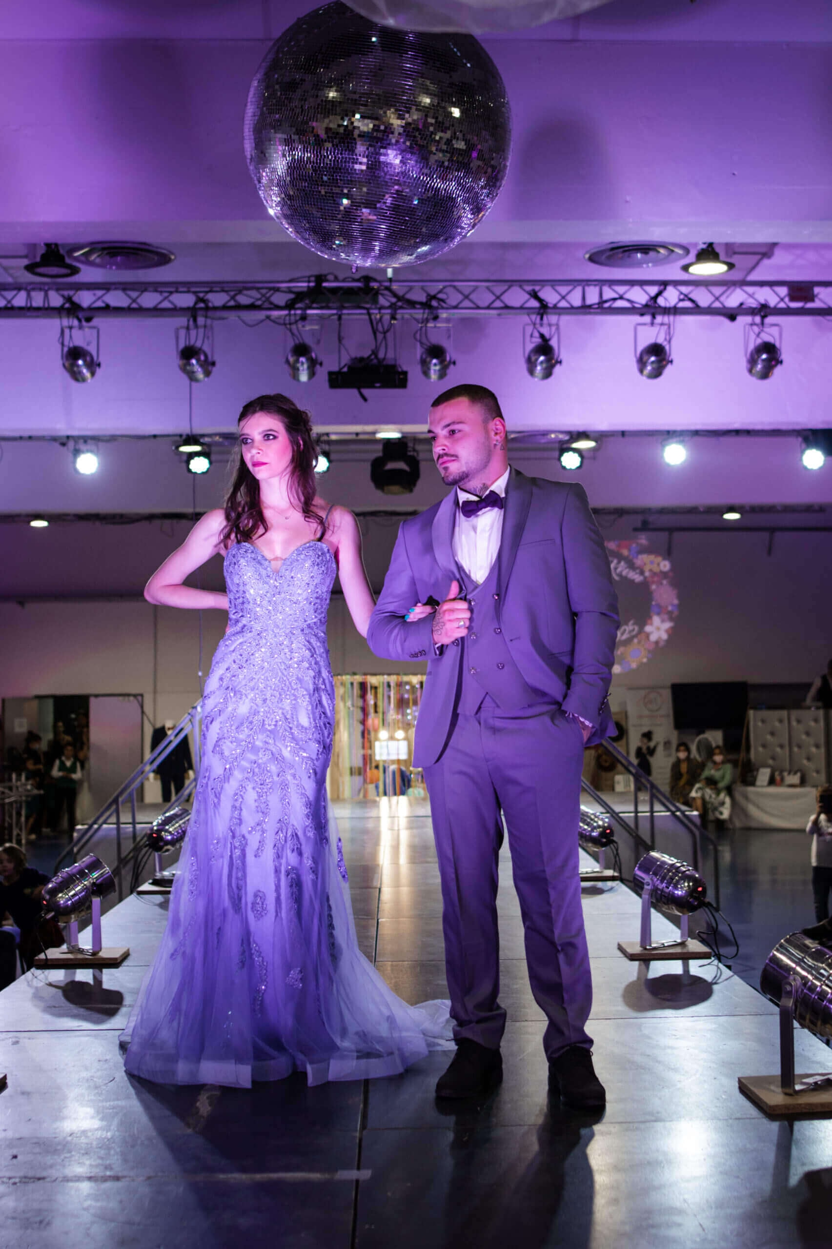 Un défilé de mode avec un modèle féminin portant une magnifique robe lavande ornée et un modèle masculin en costume gris, tous deux posant sous une boule à facettes dans une ambiance glamour.