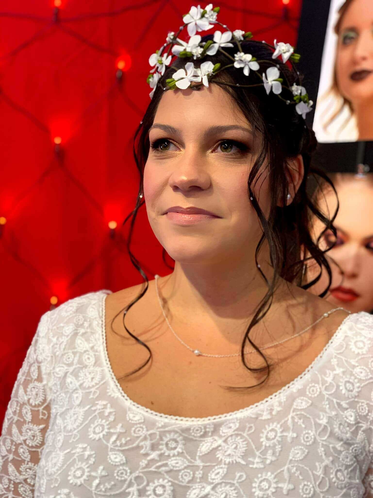 Une femme à longs cheveux bruns en ondulations pose devant un fond rouge vibrant décoré de petites lumières, vêtue d'une robe blanche brodée et d'une couronne floral composée de petites fleurs blanches, exprimant la sérénité et l'élégance.