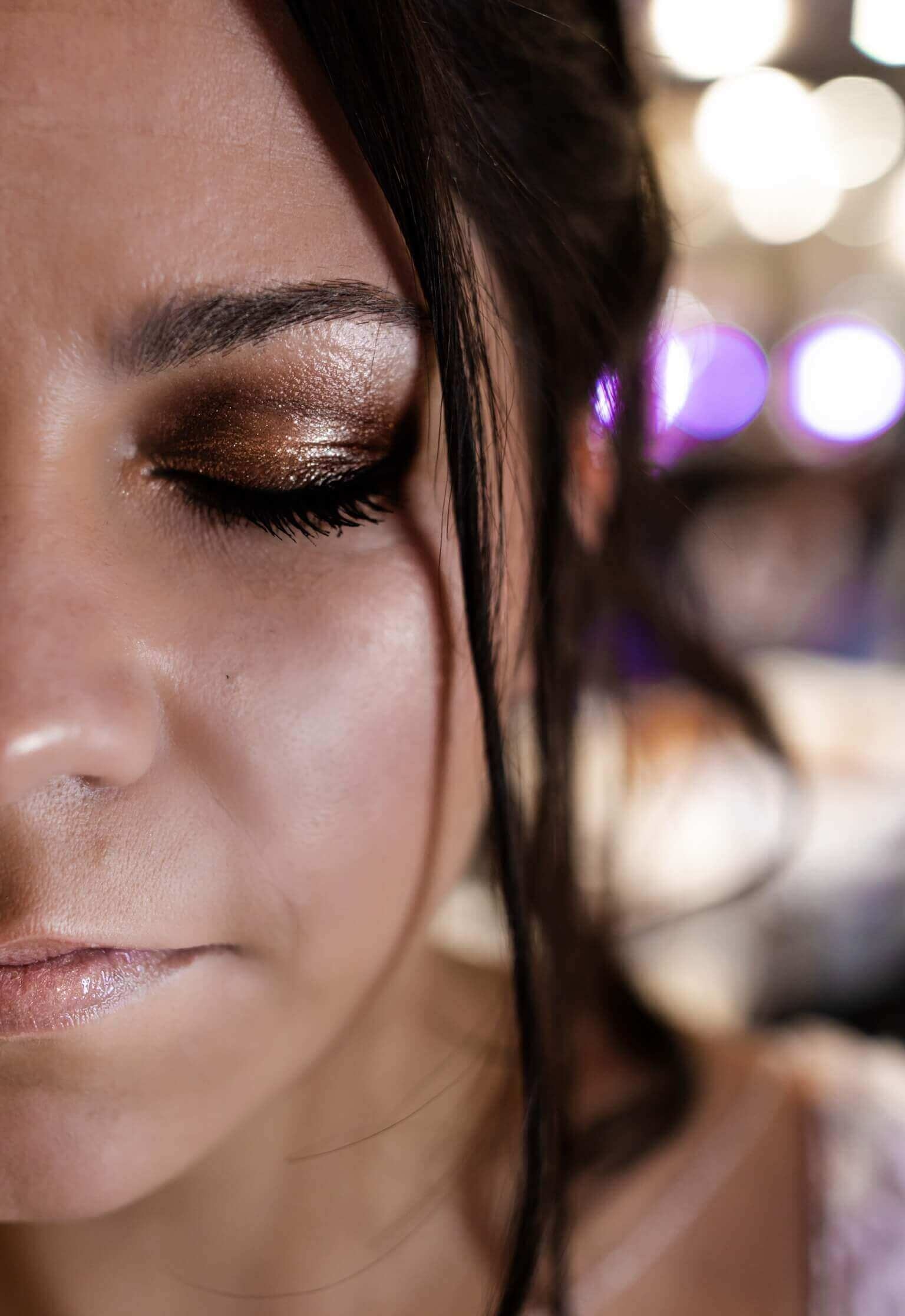 Gros plan d'une femme avec une expression sereine, mettant en valeur son maquillage avec des yeux fermés, soulignant un fard à paupières bronze scintillant et de longs cils, dans un fond flou avec un éclairage ambiant chaud.