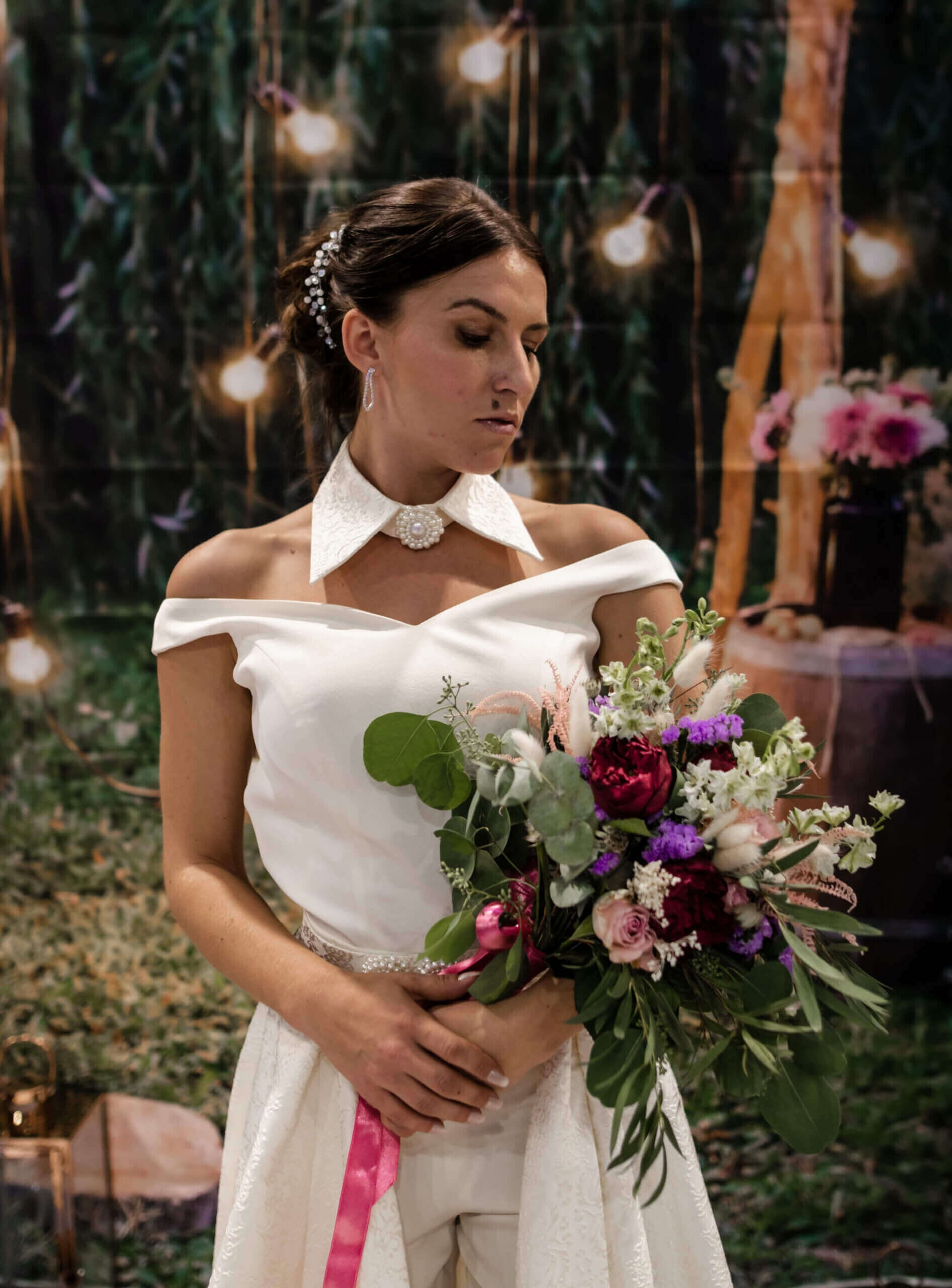 Une mariée élégante se tient gracieusement dans un cadre extérieur doucement éclairé, portant une robe de mariage off-the-shoulder avec un col délicat, tout en tenant un bouquet vibrant de nuances de rose, rouge et vert, entourée de lumières bokeh et d'éléments rustiques.