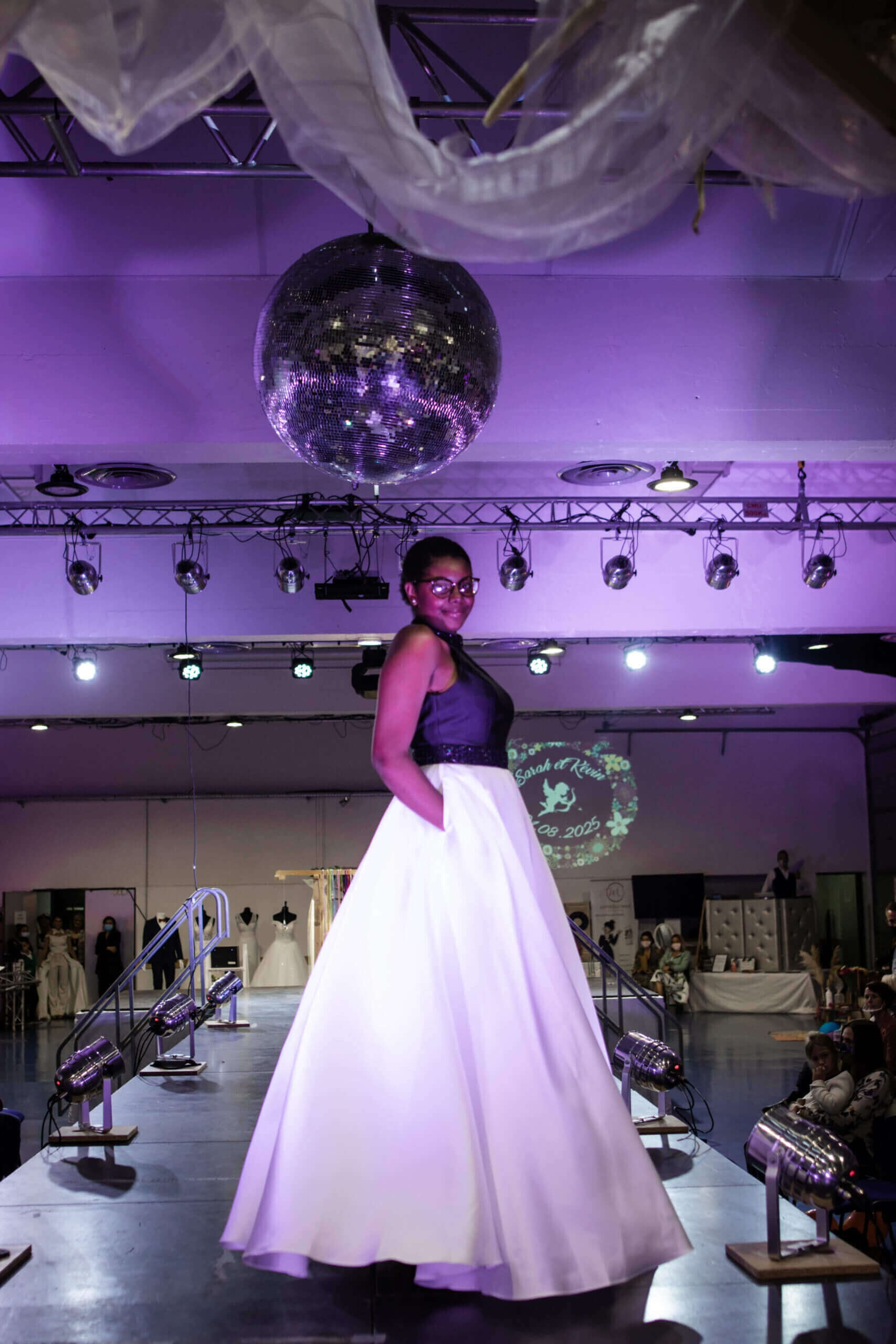 Une modèle défile sur un podium lors d'un défilé de mode, vêtue d'une longue robe sans manches noir et blanc, avec un corsage noir ajusté et une jupe blanche fluide, sous une boule à facettes qui reflète la lumière dans une ambiance festive.
