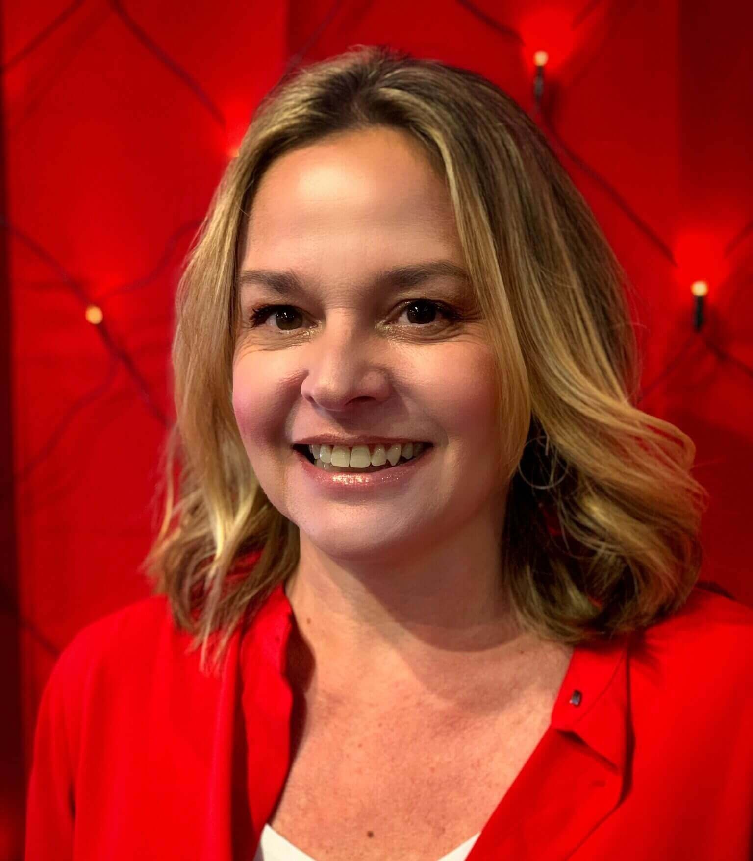 Une femme souriante devant un fond rouge vif, mettant en valeur ses cheveux blonds et sa chemise rouge.