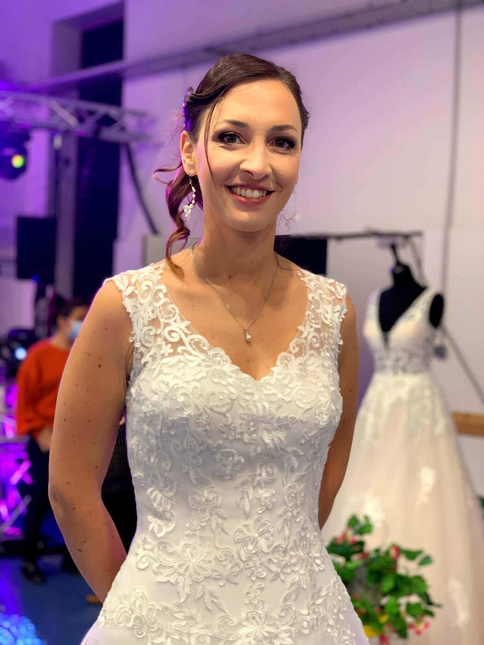 Une femme souriante portant une élégante robe de mariée blanche avec des détails en dentelle, dans une ambiance de célébration, avec un arrière-plan flou de silhouettes de personnes et d'autres robes.