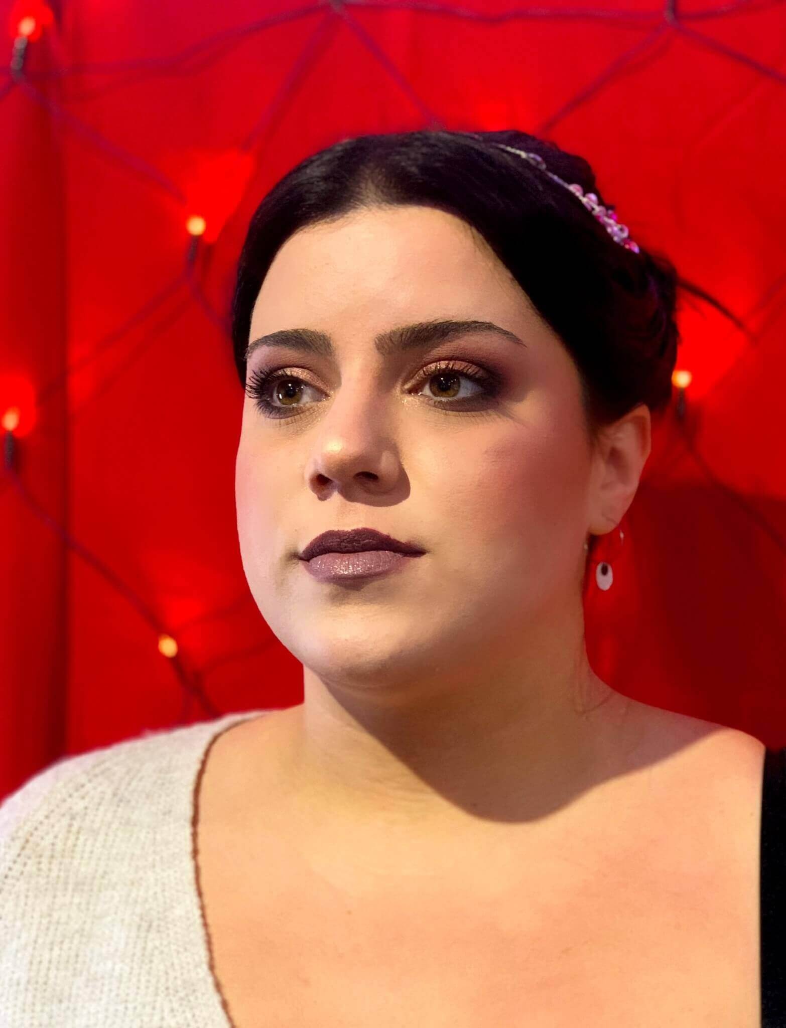 Femme aux cheveux foncés, coiffée élégamment, portant un pull léger avec un maquillage subtil mettant en valeur ses yeux et lèvres brillants devant un fond rouge décoré de lumières douces.