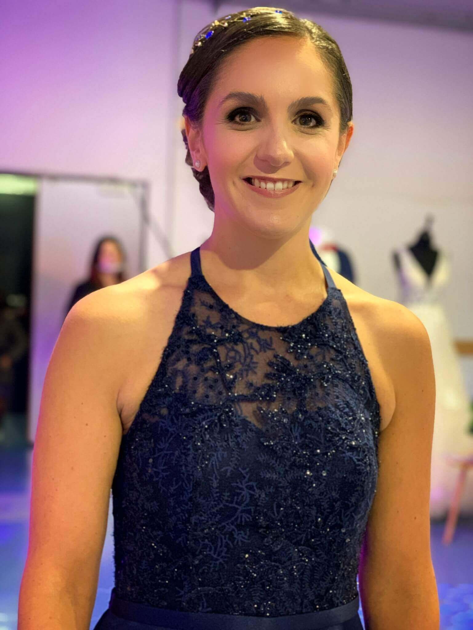 Une femme souriante regardant directement l'appareil photo, vêtue d'une élégante robe bleu foncé avec des détails en dentelle et des perles sur le corsage, dans un espace tamisé lors d'un événement de mode.