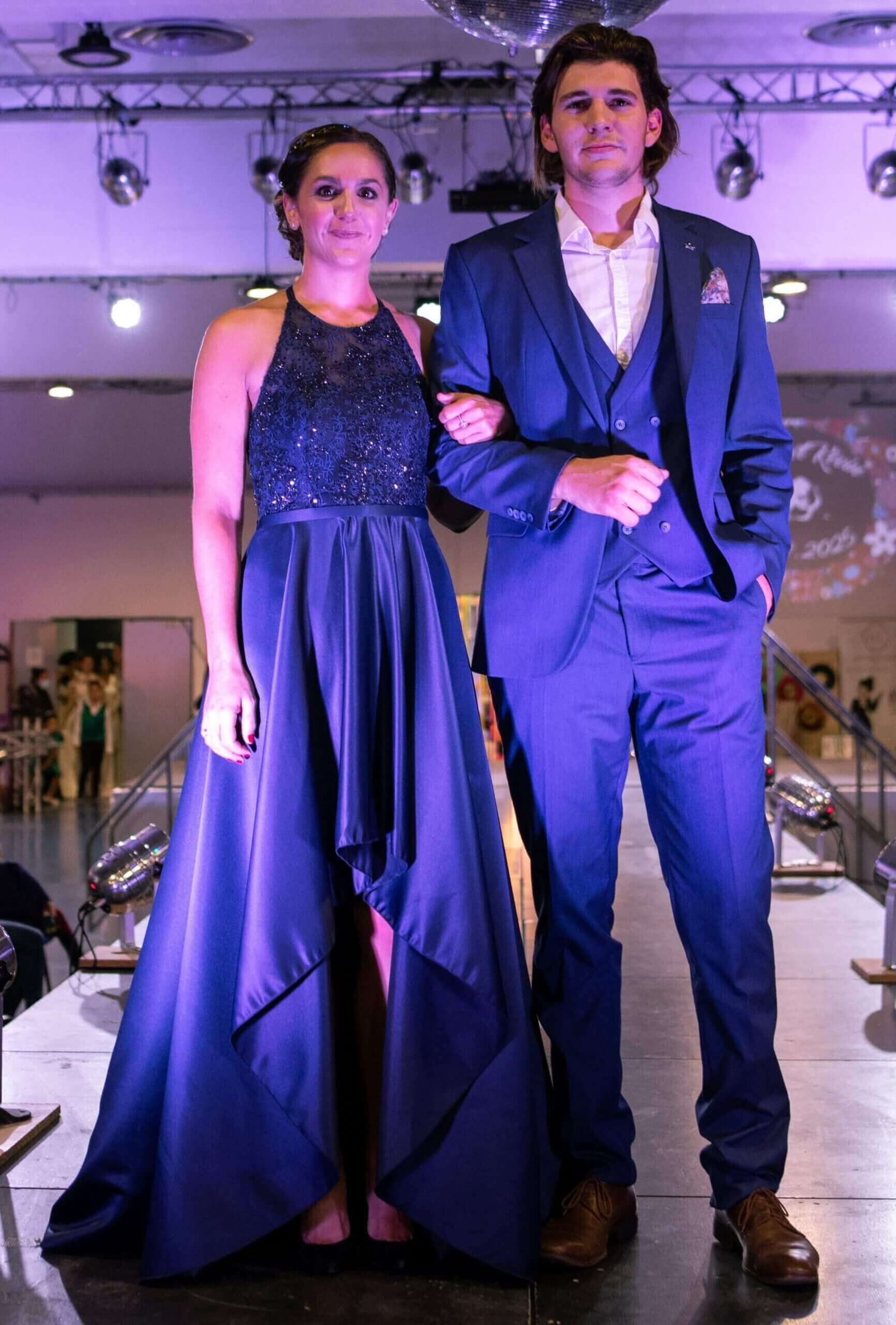 Un homme et une femme posent ensemble sur un podium de mode, la femme portant une élégante robe de soirée bleue nuit tandis que l'homme est habillé d'un costume bien ajusté. L'éclairage scintillant et un disco ball en arrière-plan ajoutent une ambiance glamour à cet événement de mode.