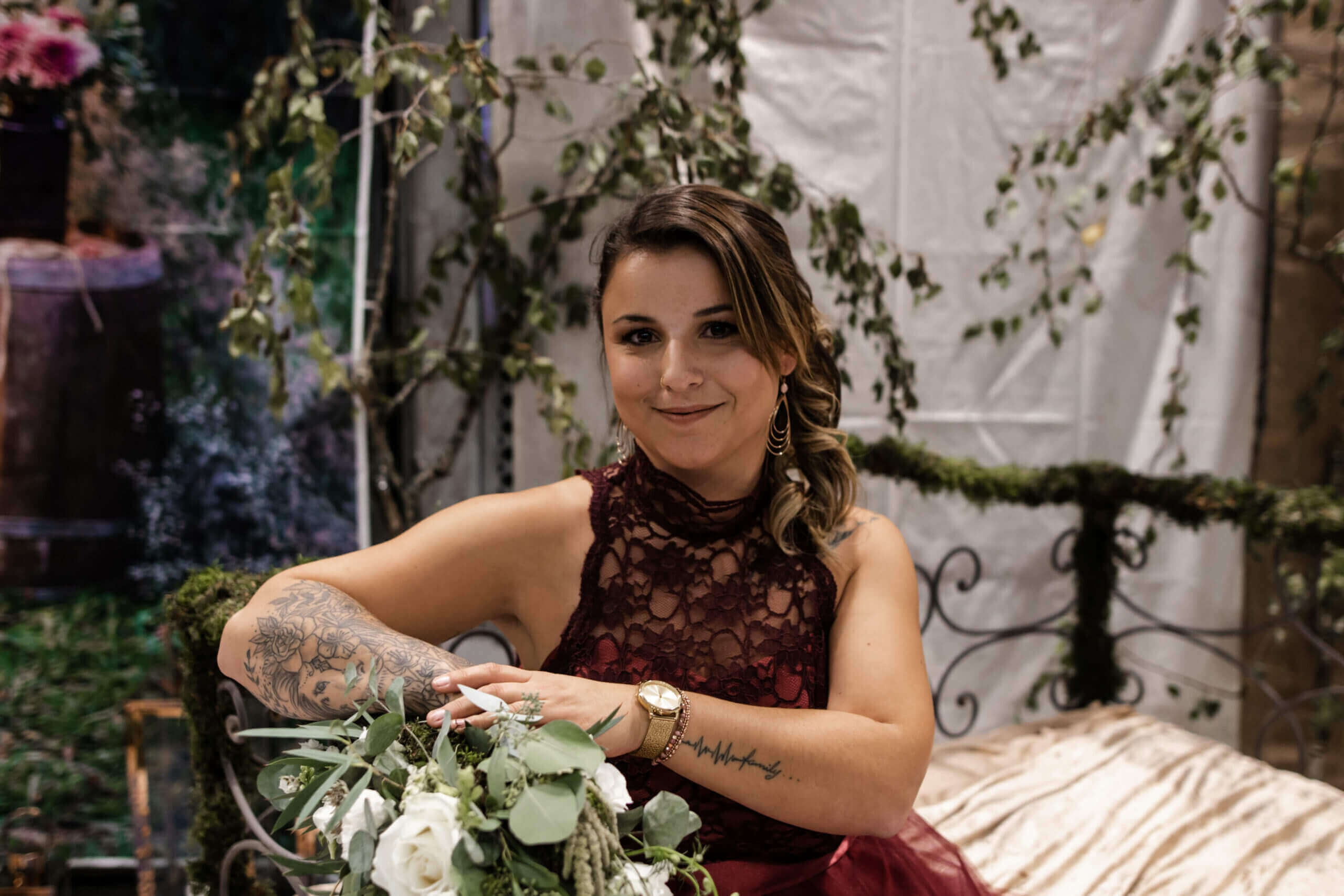 Une jeune femme aux longs cheveux ondulés sourit à l'appareil photo, vêtue d'une robe bordeaux en dentelle et portant des bijoux, tout en tenant un bouquet de fleurs blanches et de verdure dans un cadre naturel et intime.